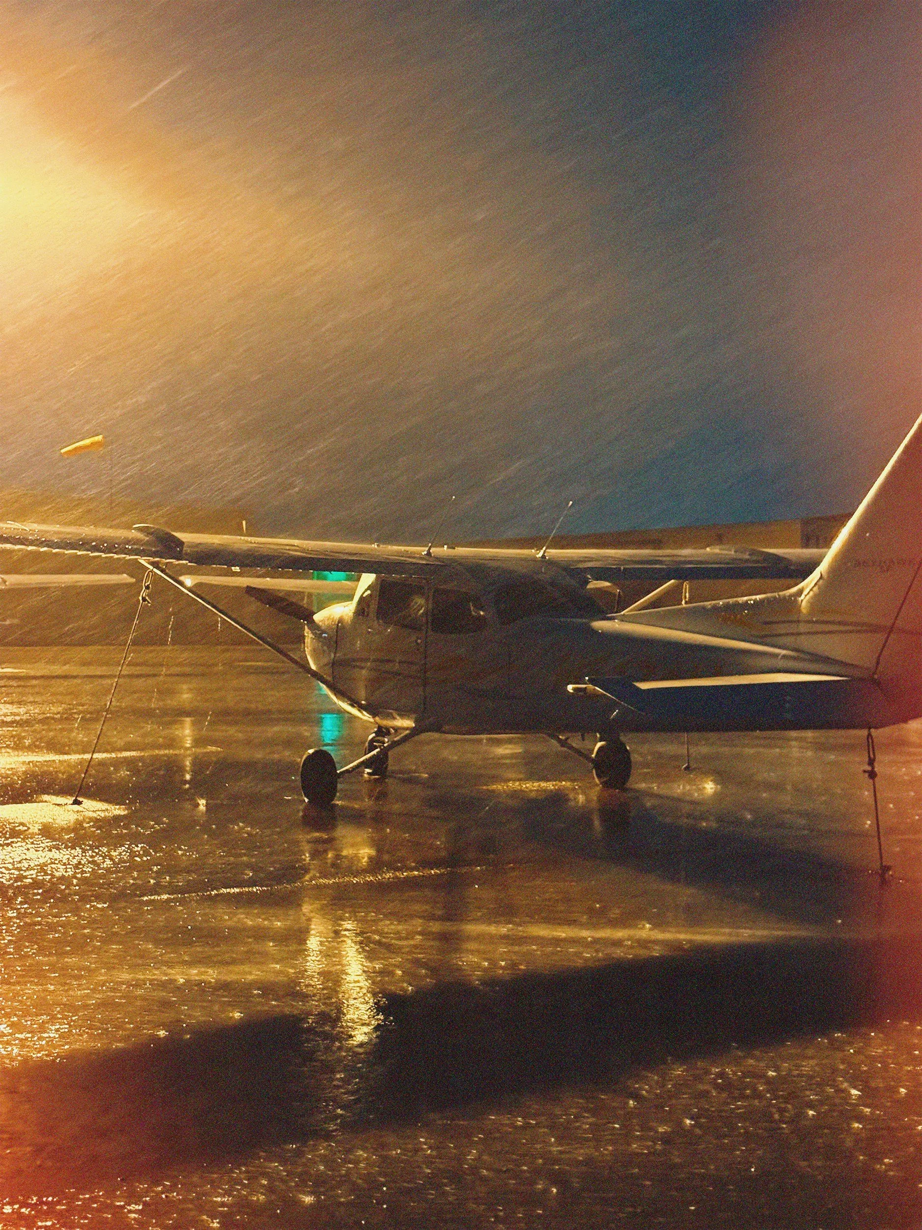 Small propeller airplane on a wet tarmac at night, illuminated by a nearby streetlight, with a dark cloudy sky overhead.