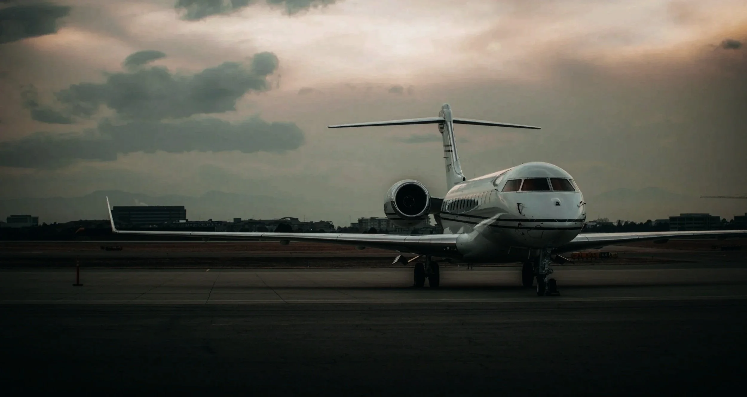 Private jet airplane parked on the runway under an overcast sky with city buildings and mountains in the background.
