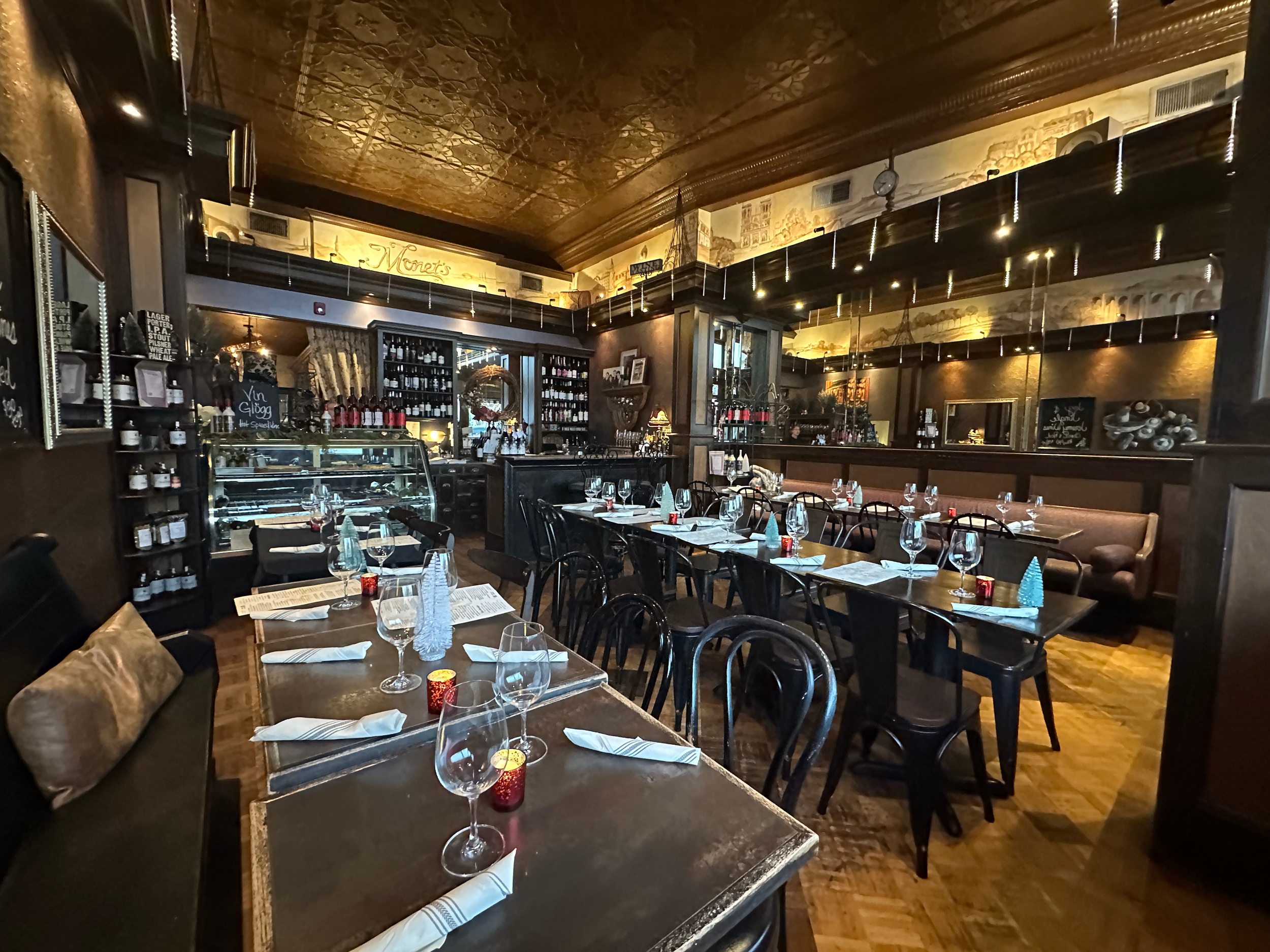 Interior of a cozy, rustic restaurant with wooden tables set with wine glasses, napkins, and small candles, decorated with holiday-themed items, and a bar with shelves of bottles in the background.