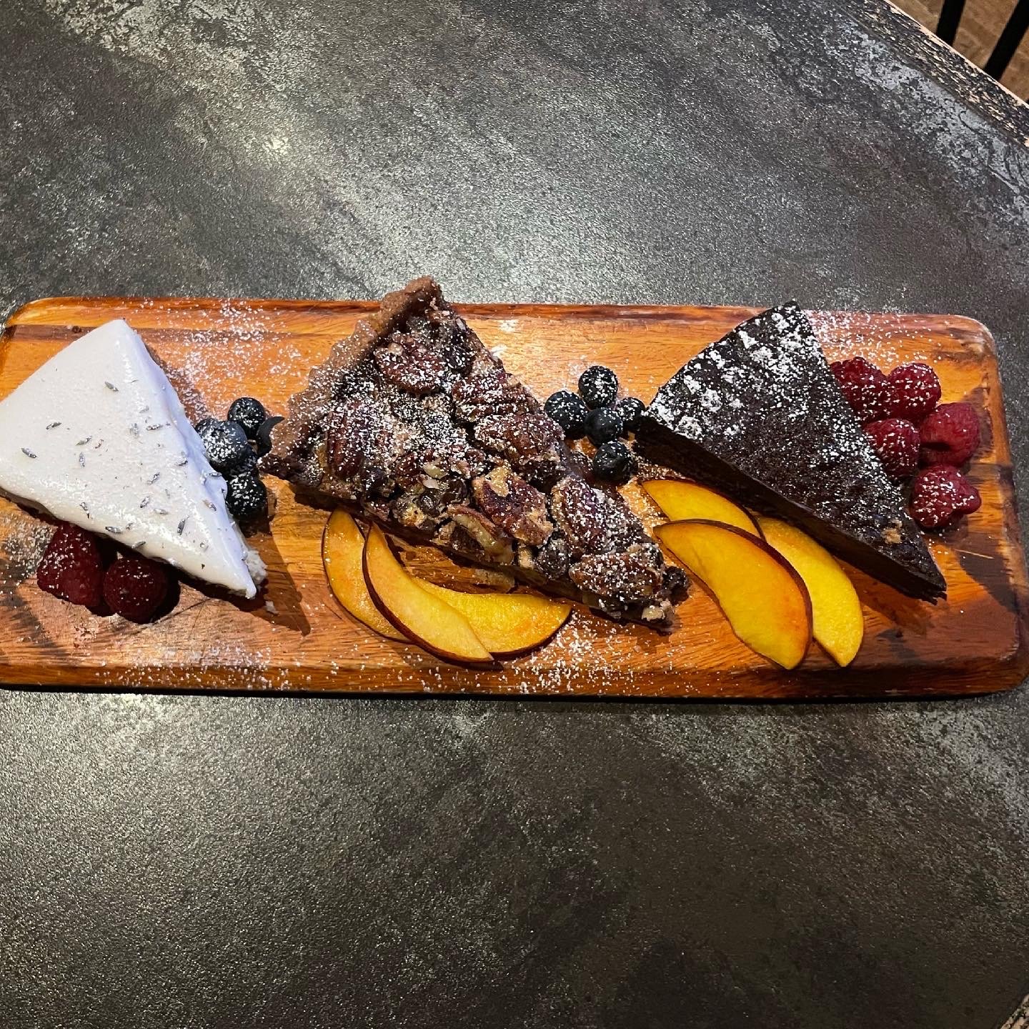A wooden platter with three slices of different artisan cheeses, fresh berries, and sliced peaches, dusted with powdered sugar.