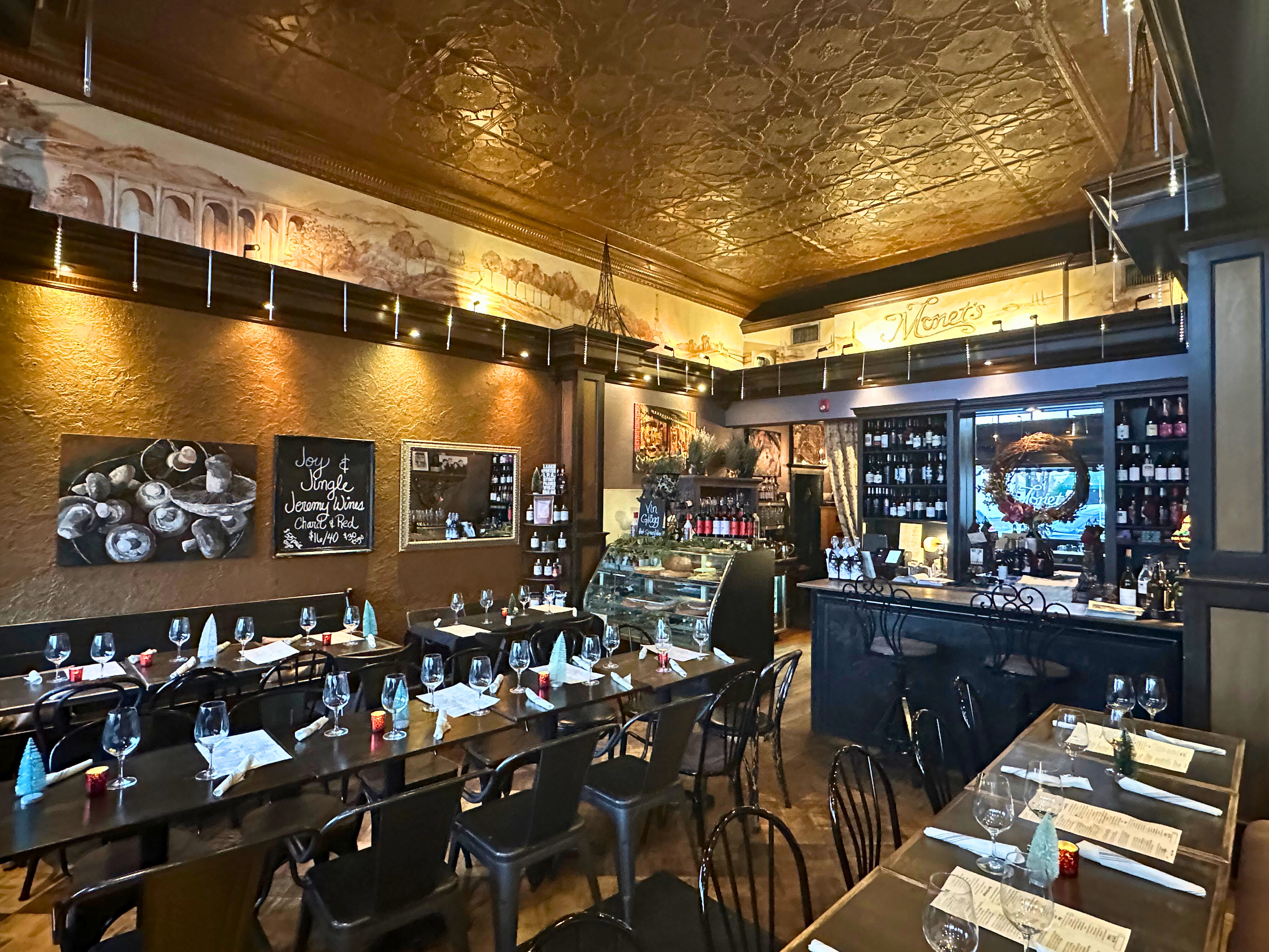 Cozy restaurant interior with wooden tables set with wine glasses, napkins, and candles, decorated with paintings, a chalkboard menu, and wine bottles on shelves, featuring warm lighting and elegant decor.