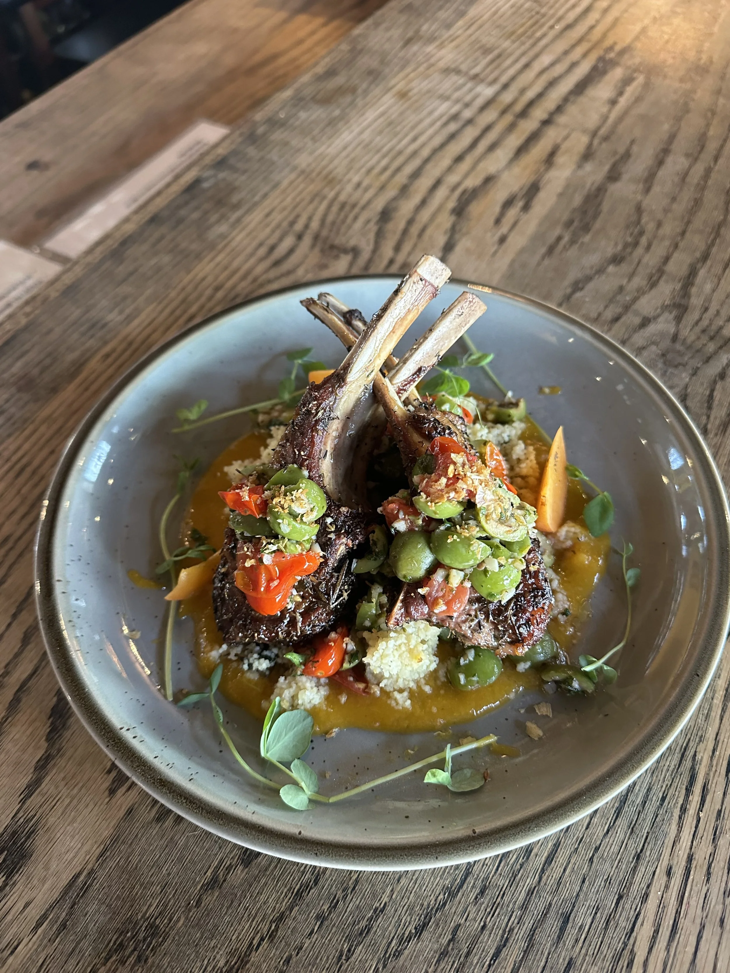 Gourmet dish featuring two grilled lamb chops topped with green peas and diced red peppers, garnished with microgreens, served on a bed of yellow sauce on a gray ceramic plate.