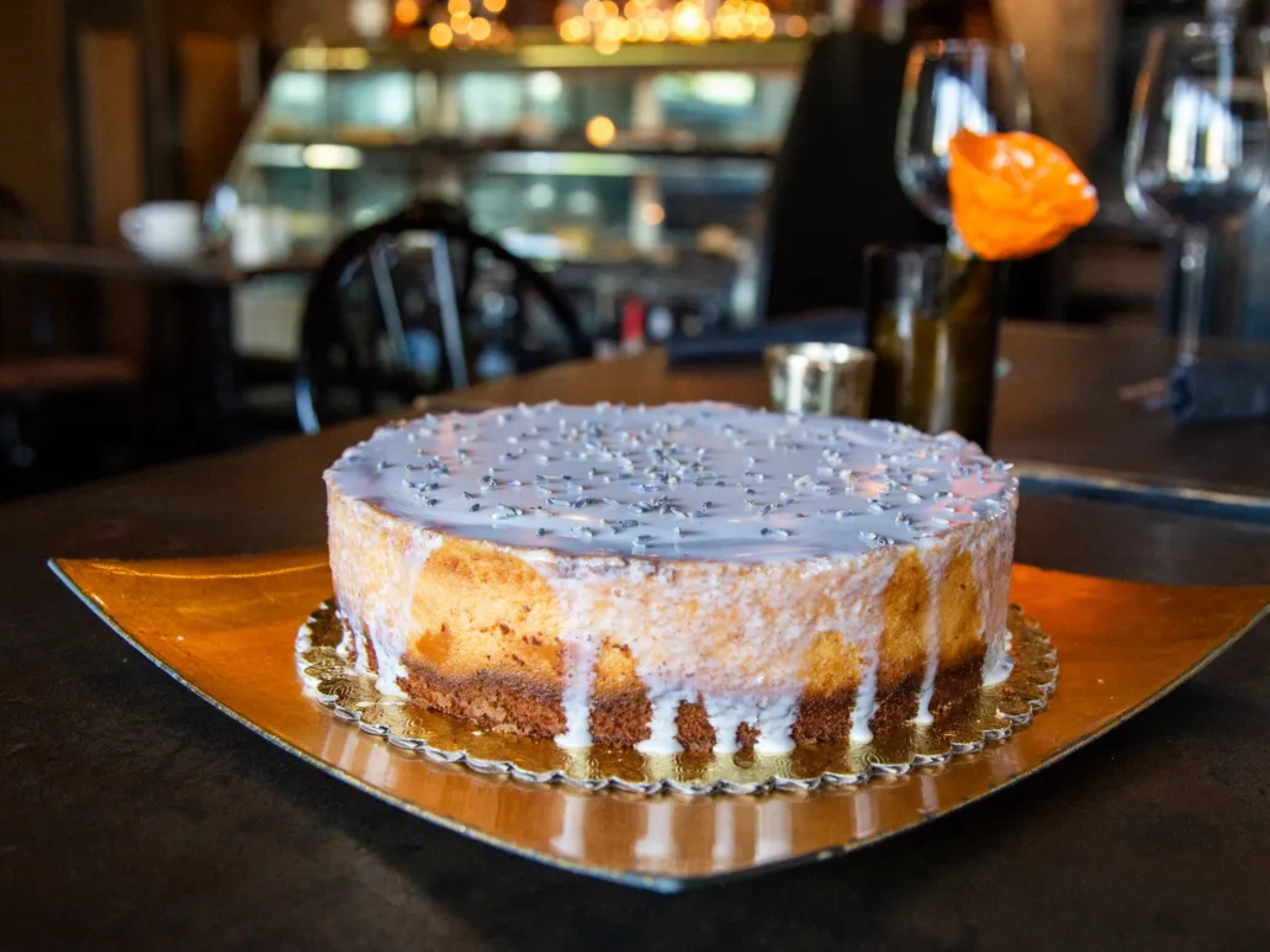 A round layered cake with white icing and sprinkles on top, sitting on a gold-colored tray in a restaurant setting.