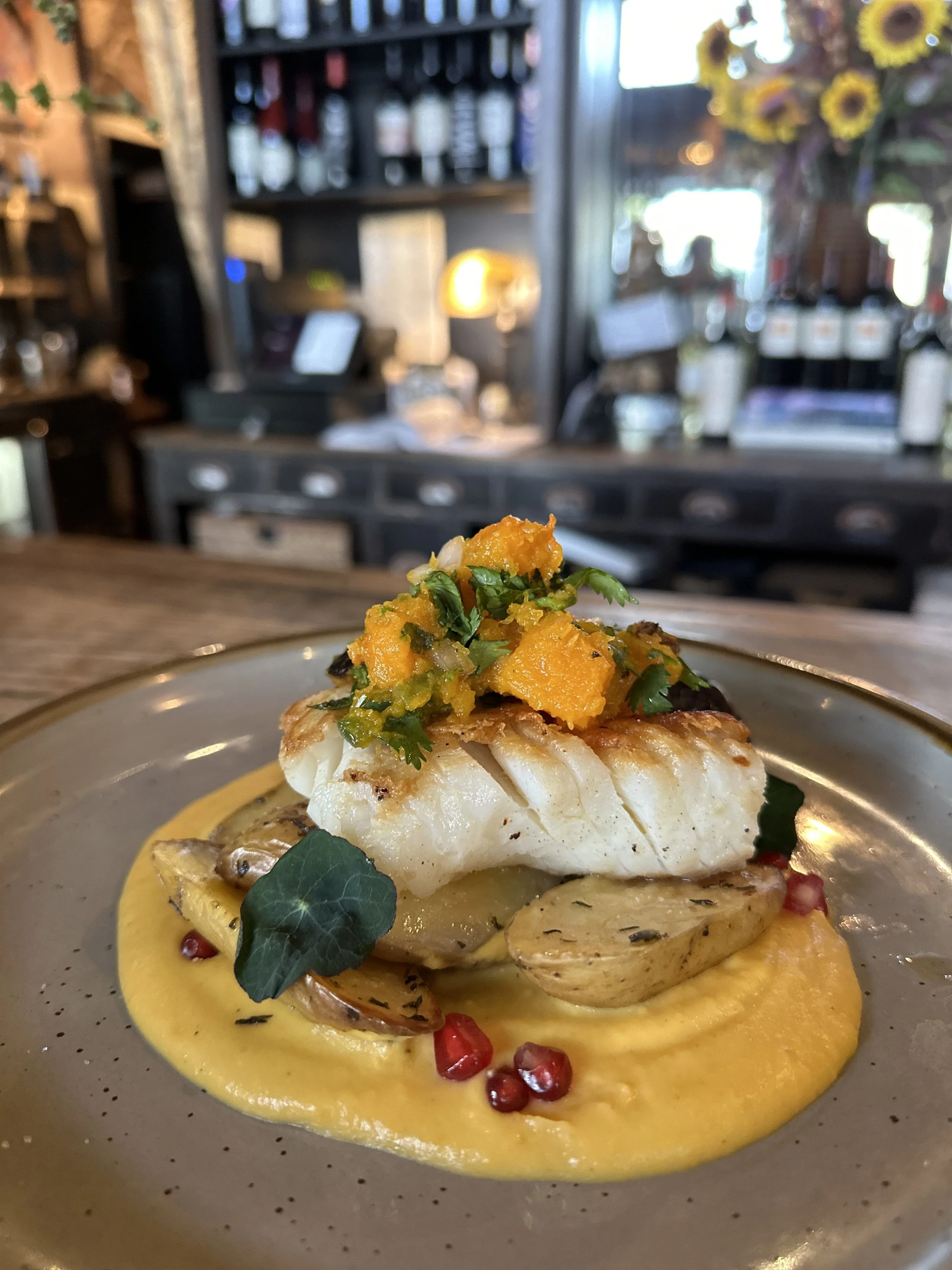 A plated dish featuring a white fish fillet topped with a yellow and green vegetable mixture, served on a bed of creamy yellow sauce with roasted potatoes, pomegranate seeds, and a green leaf garnish, set in a restaurant with warm lighting and a blur