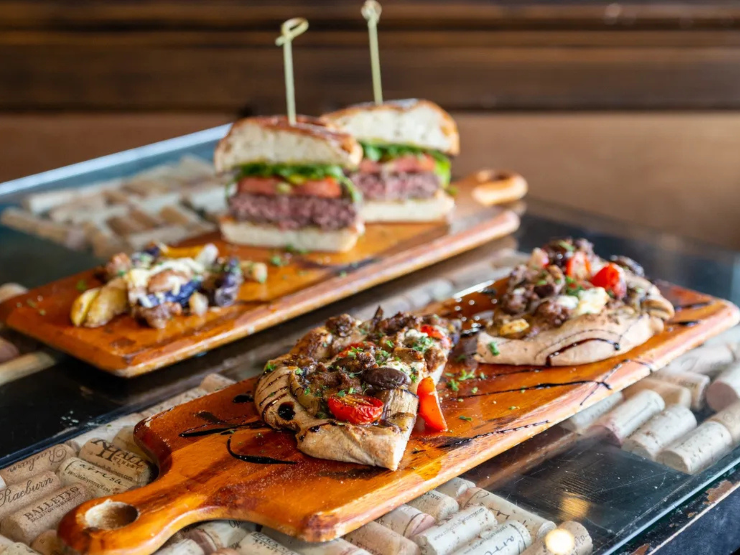Two bruschetta with vegetables and cheese, and a cheeseburger with lettuce, tomato, and beef patty on a wooden board.