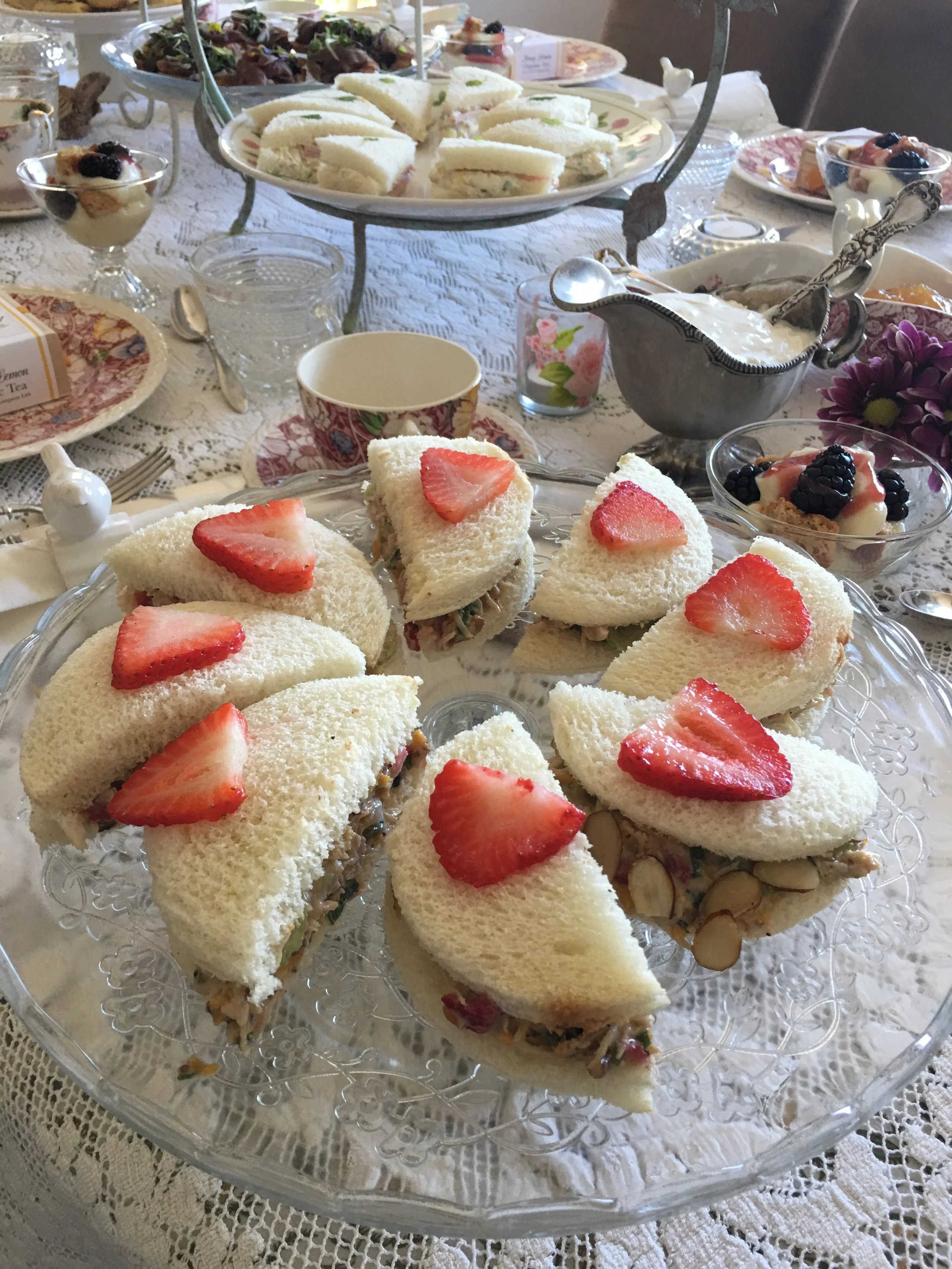 Plate of finger sandwiches with strawberries on top, assorted desserts, and a tiered tray of more sandwiches on a lace tablecloth.