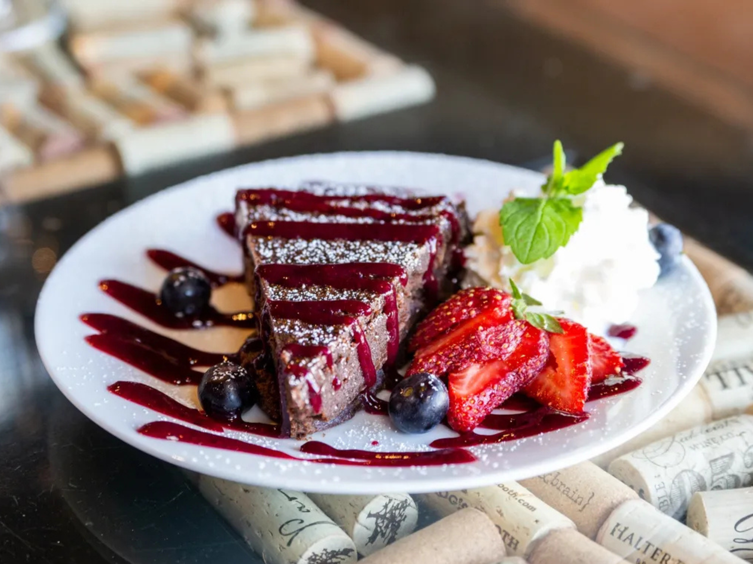 Chocolate cake slices with berry sauce, fresh strawberries, blueberries, whipped cream with a mint leaf, served on a white plate decorated with berry sauce and powdered sugar.