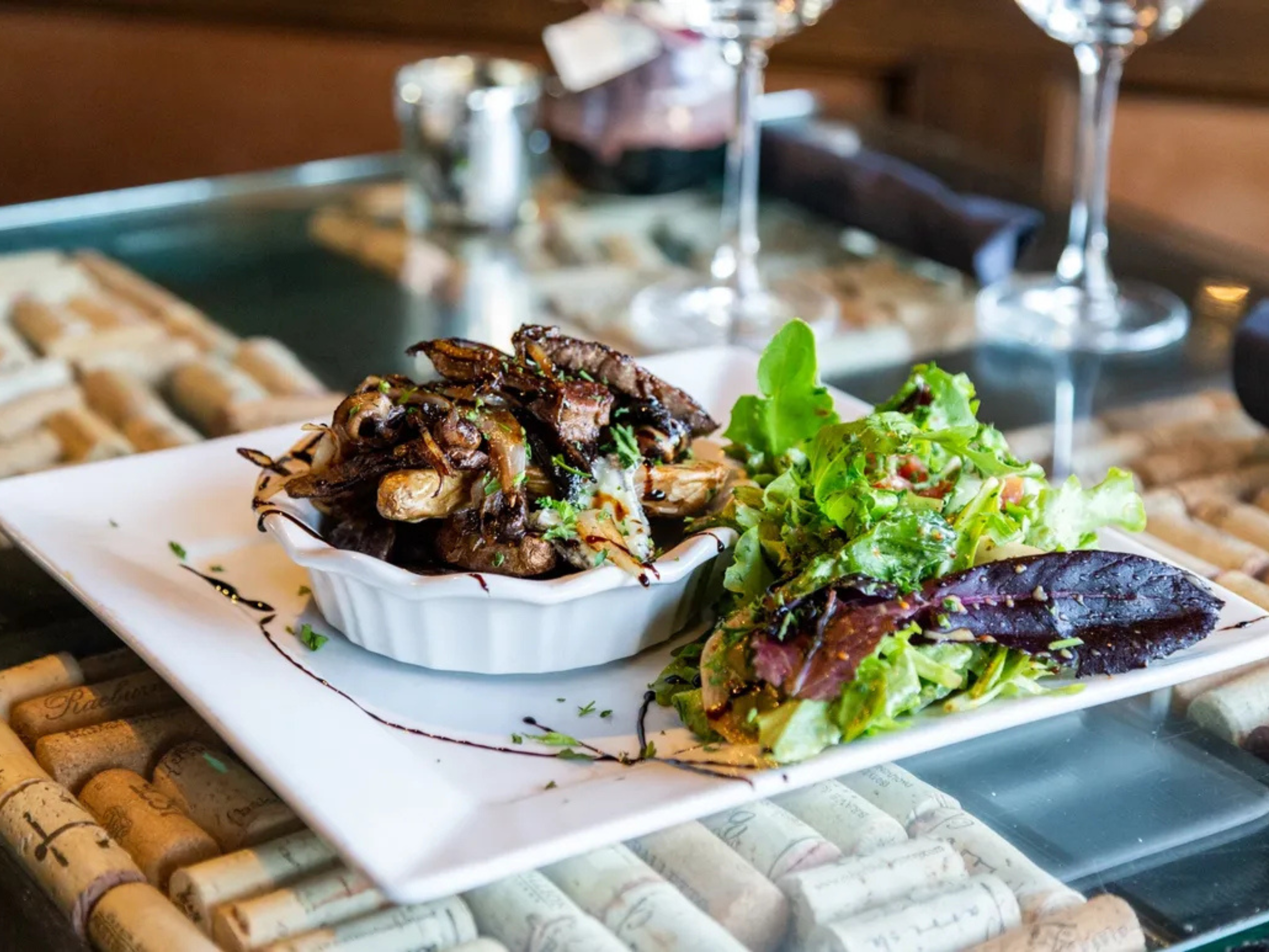 A white rectangular plate with a small dish of cooked mushrooms and onions, a side salad with mixed greens, and a leaf of red lettuce. The plate is on a table with wine glasses and wine bottles in the background.