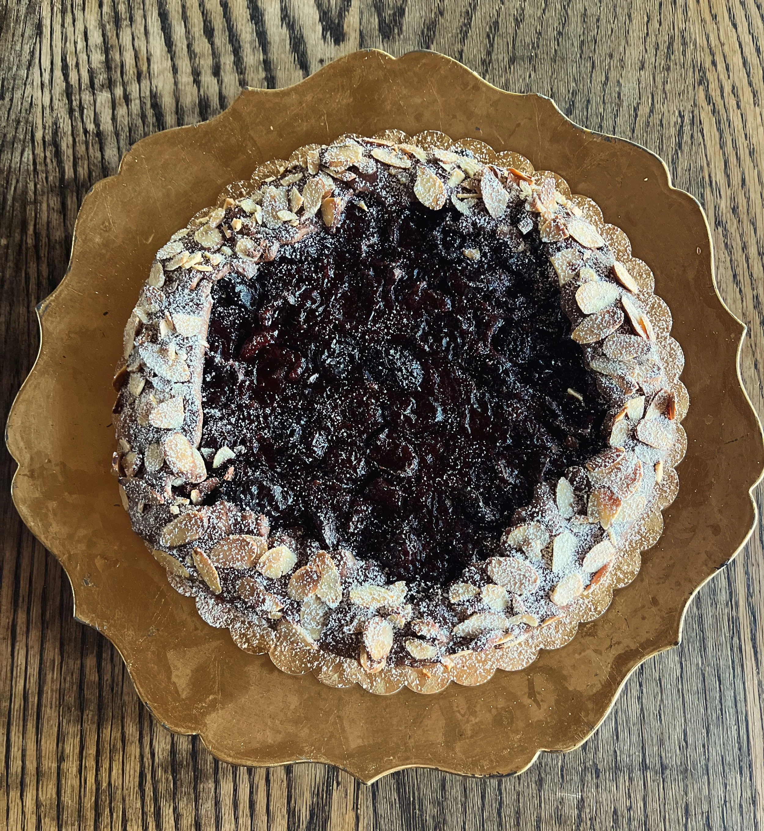 A round fruit tart with a dark berry filling, topped with sliced almonds and powdered sugar, on a gold scalloped edge plate on a wooden surface.