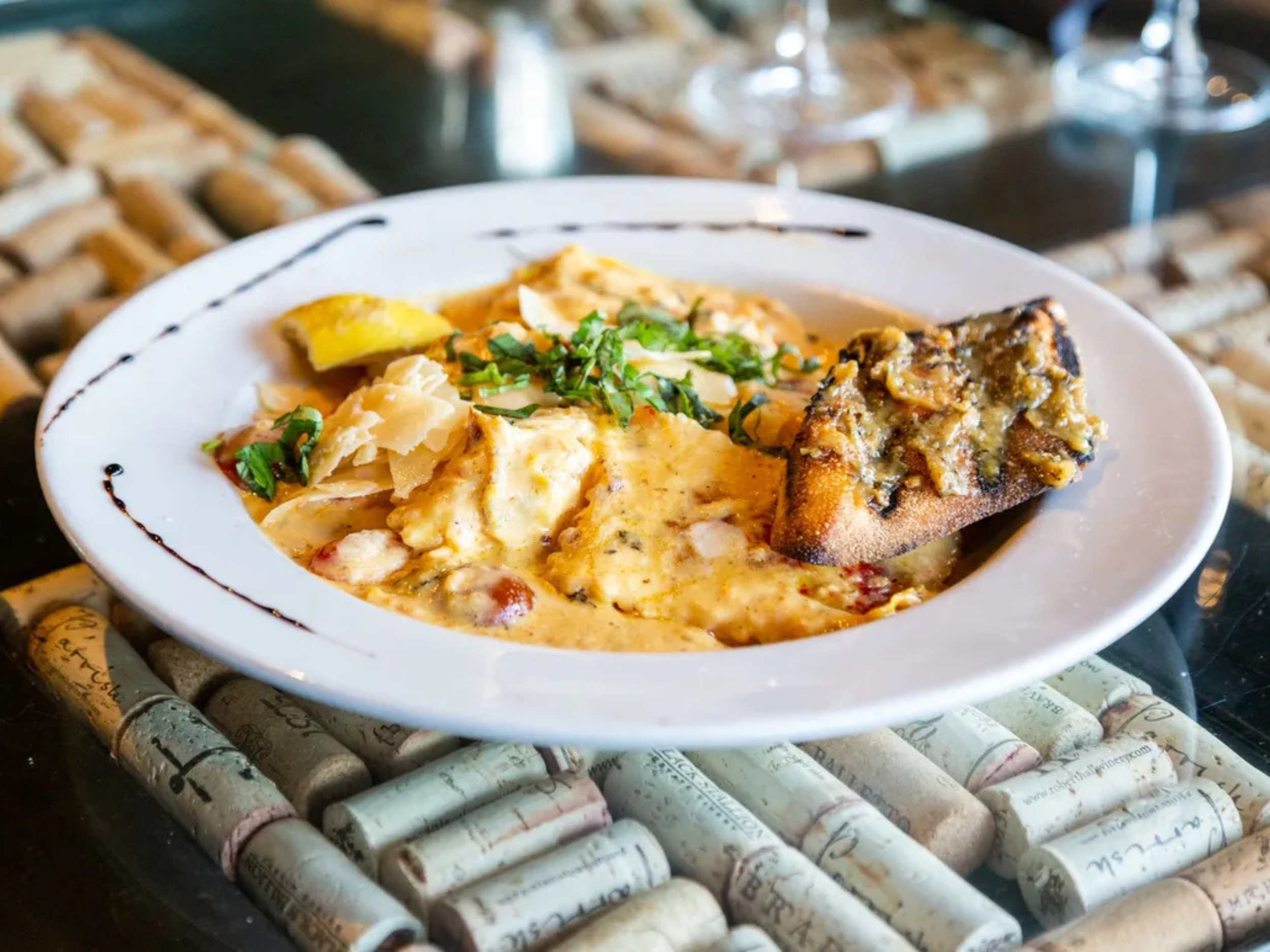Dish with melted cheese, herbs, bread topped with a mixture, potato, and sliced lemon on a white plate with a black decorative border.