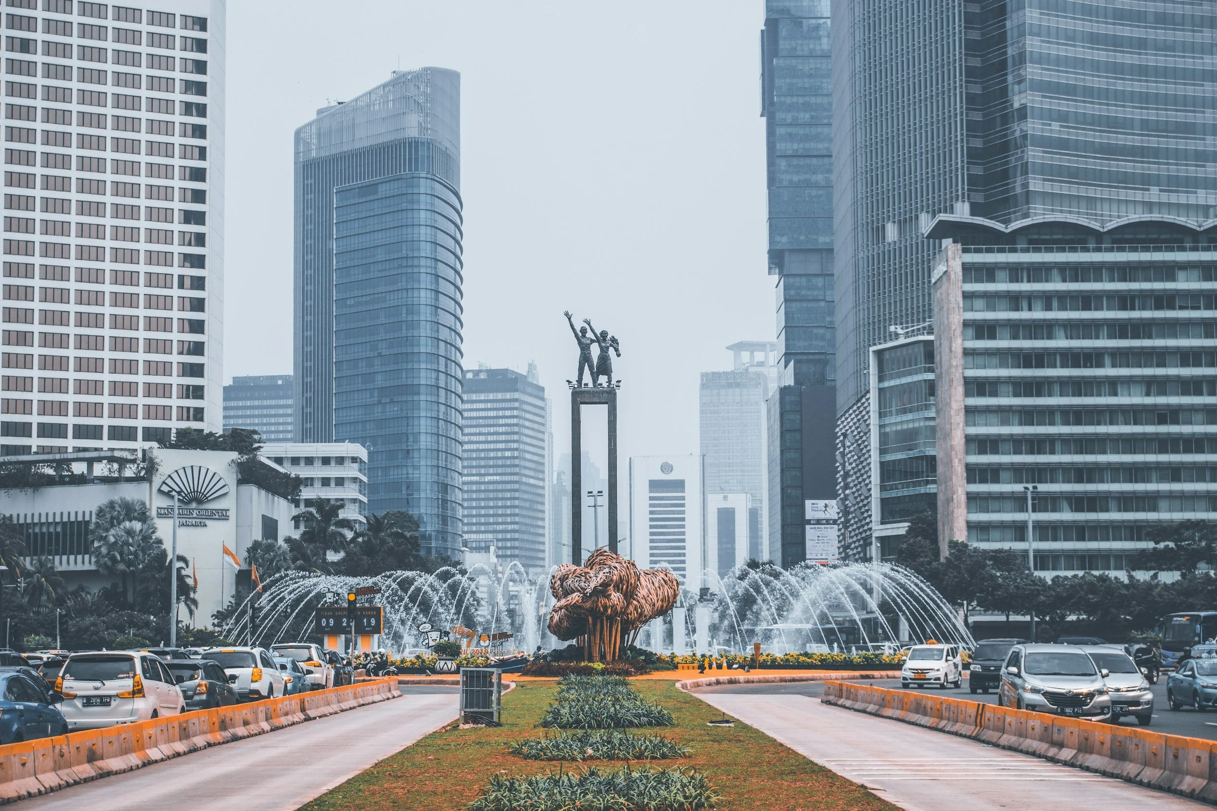 City street with tall modern skyscrapers, a fountain with a sculpture of two people, and a parking area with cars in the foreground.