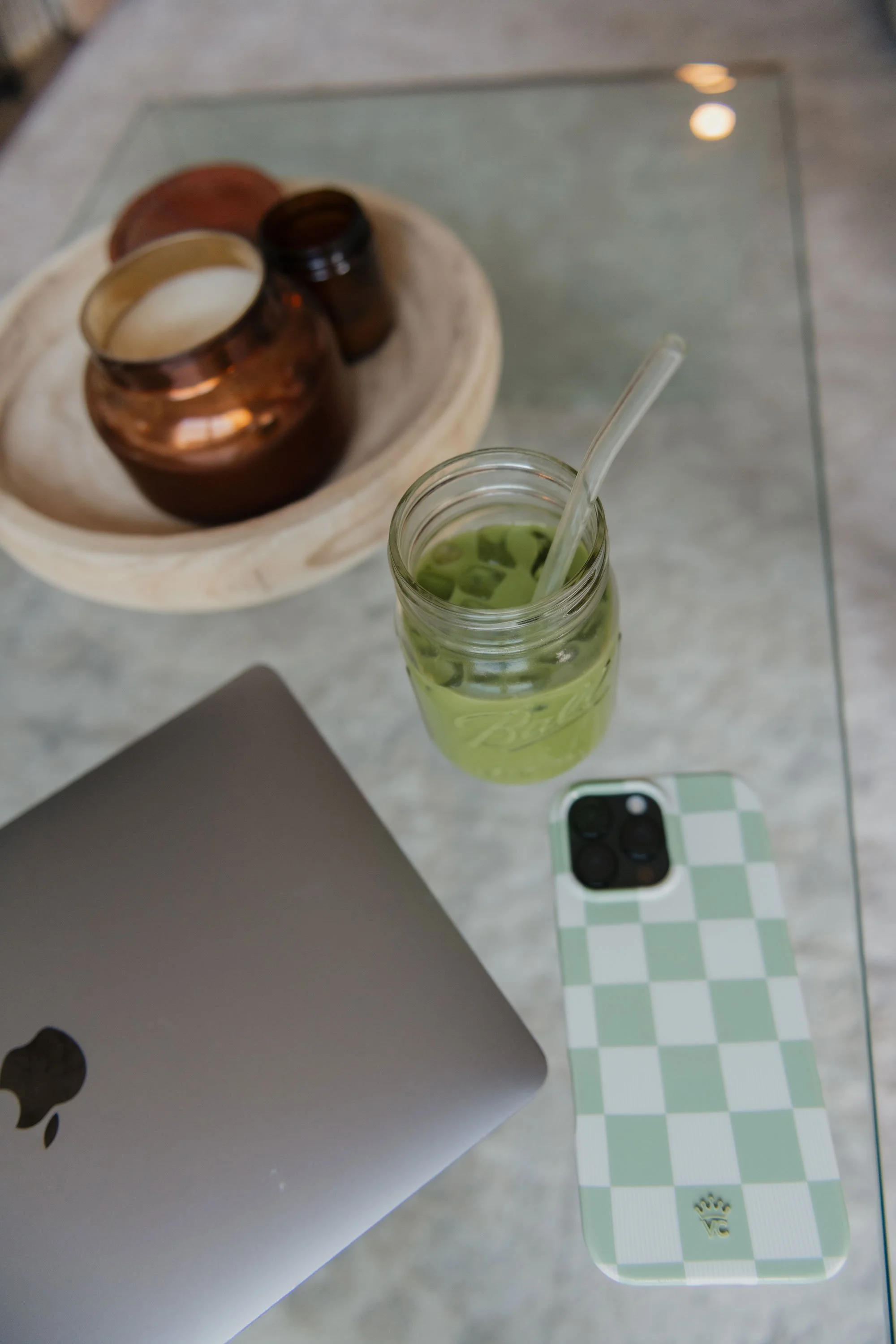 A glass jar with green tea and ice, a smartphone with a checkered green and white case, a closed laptop, and a decorative tray with jars in a cozy setting.