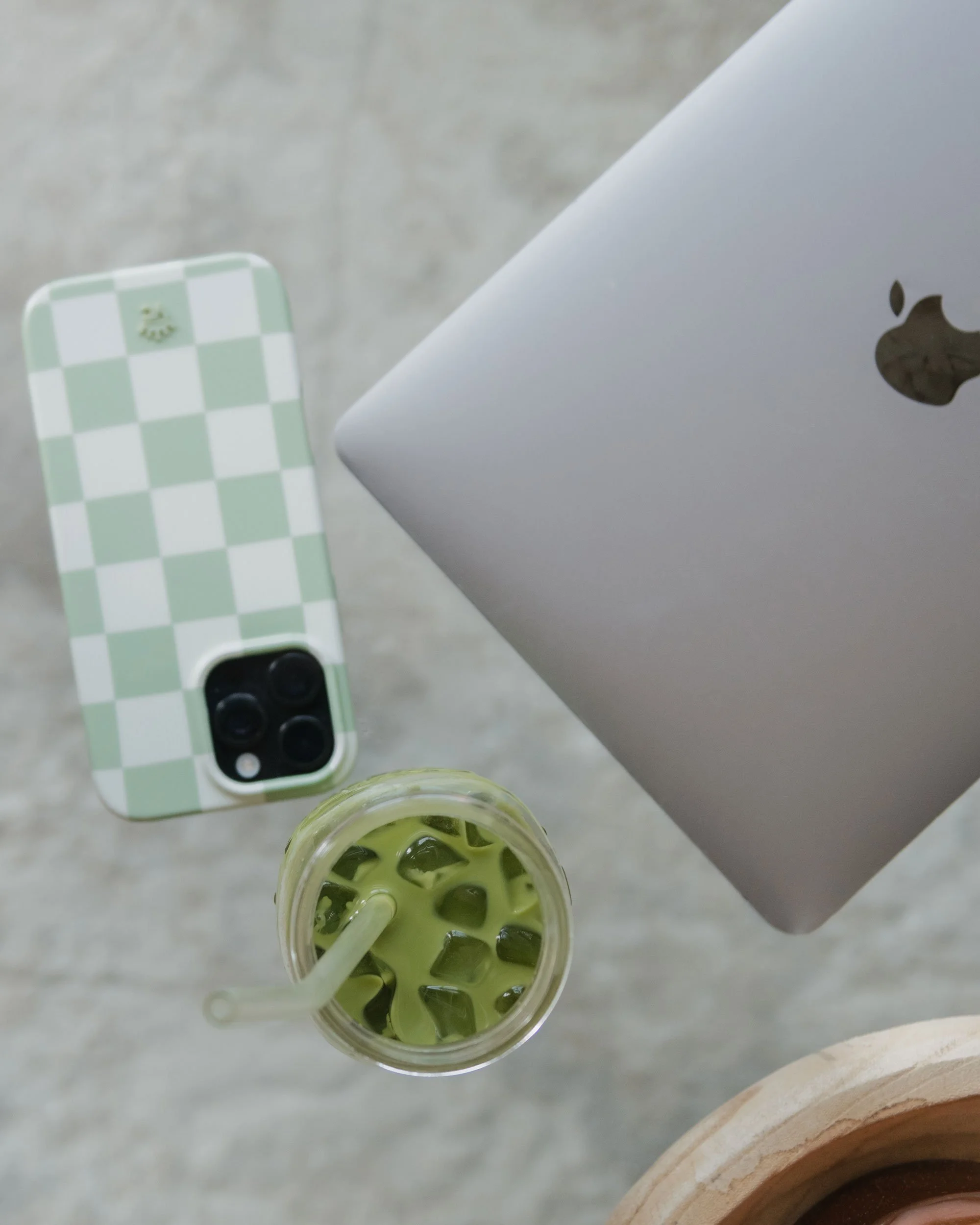 Overhead view of a MacBook, a green checkered phone case with a small frog detail, and a drink with ice and green liquid with a straw on a gray surface.