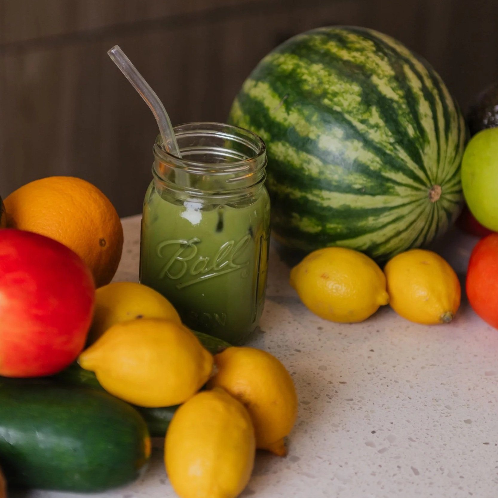A collection of fresh fruits including apples, lemons, oranges, a watermelon, and a cucumber on a kitchen counter, with a mason jar of green juice with a straw among them.