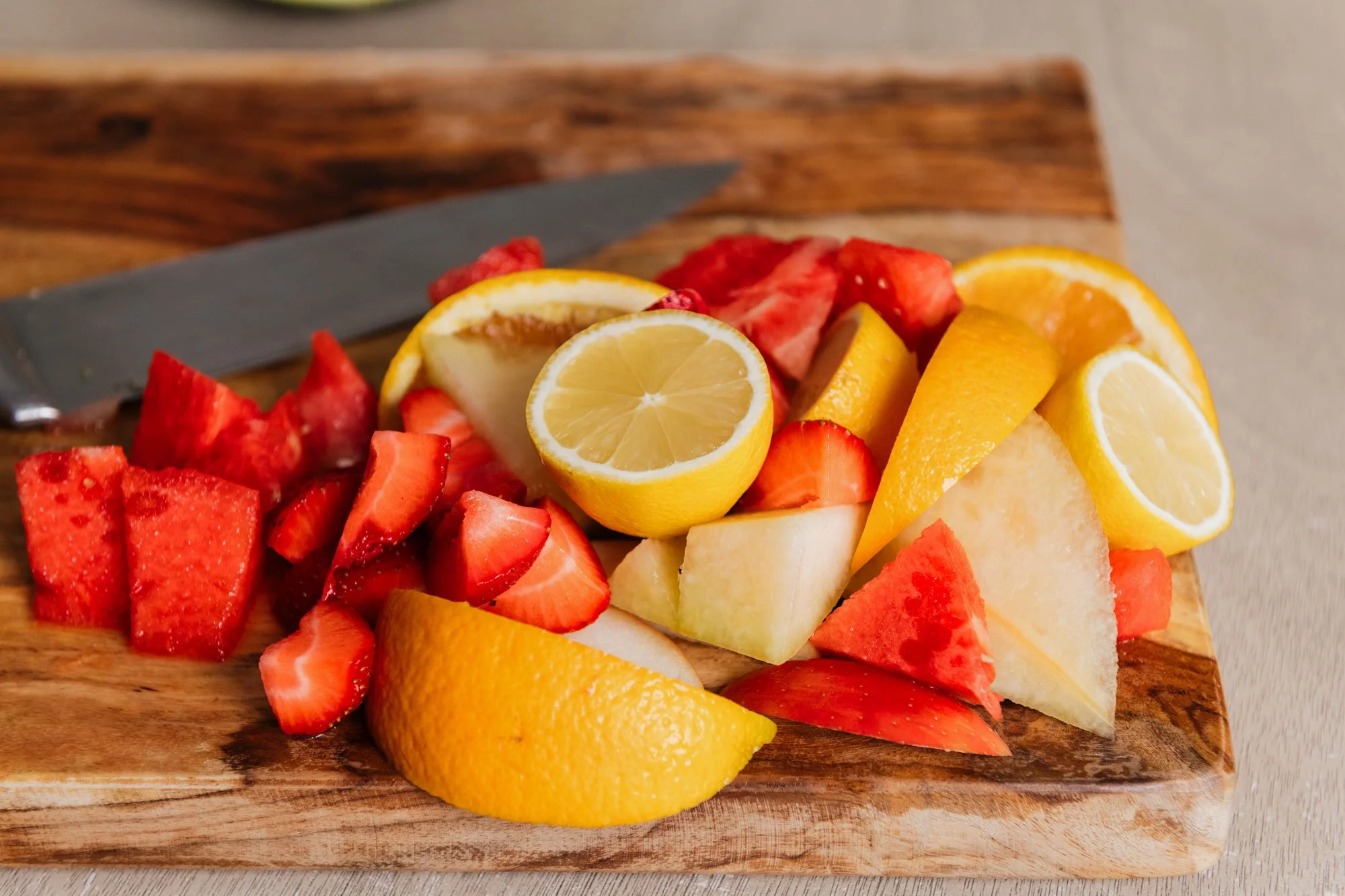 Assorted sliced fruits including strawberries, lemons, oranges, and cantaloupe on a wooden cutting board with a knife.