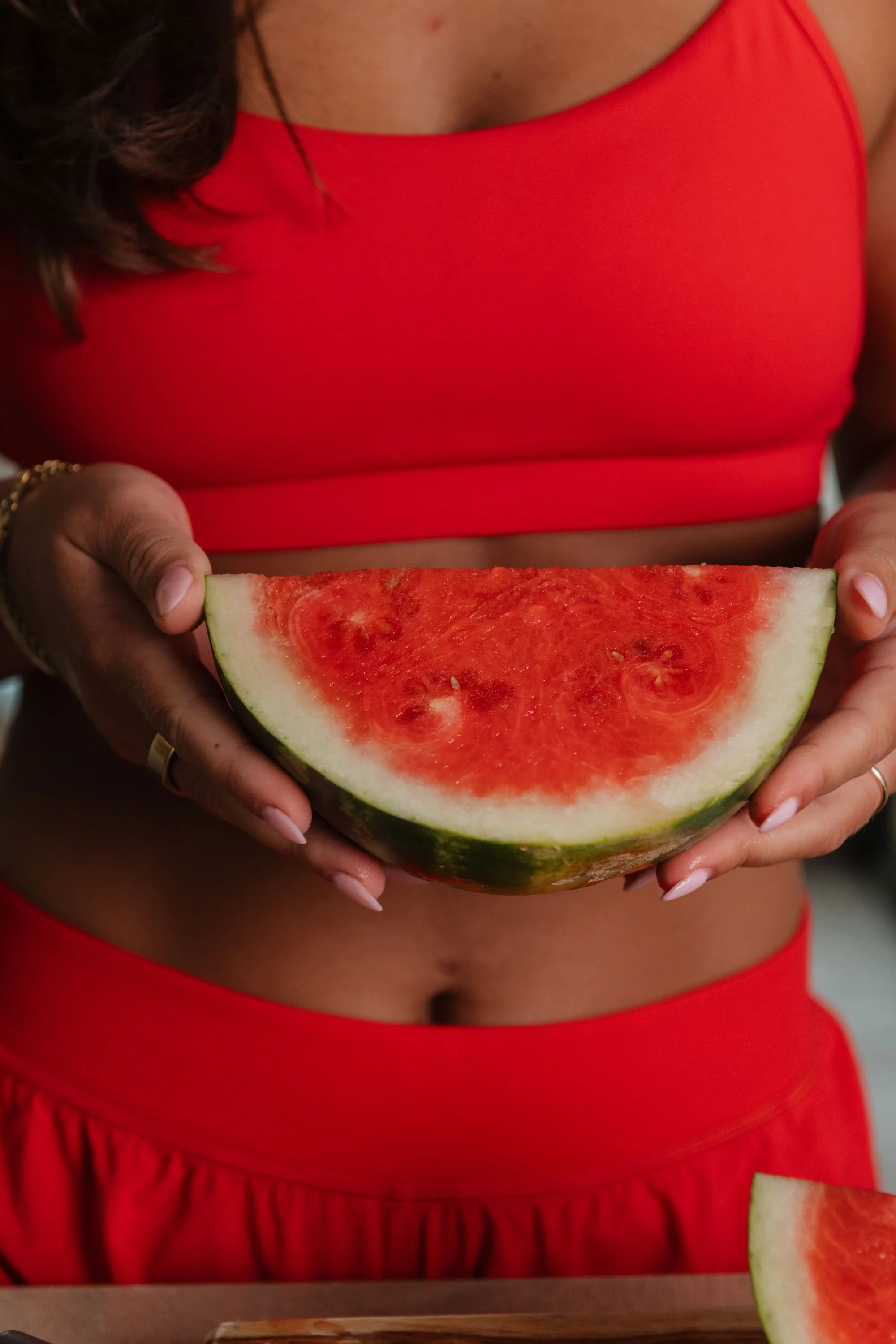 A person in a red tank top and red skirt holding a slice of watermelon with vibrant red flesh and green rind.