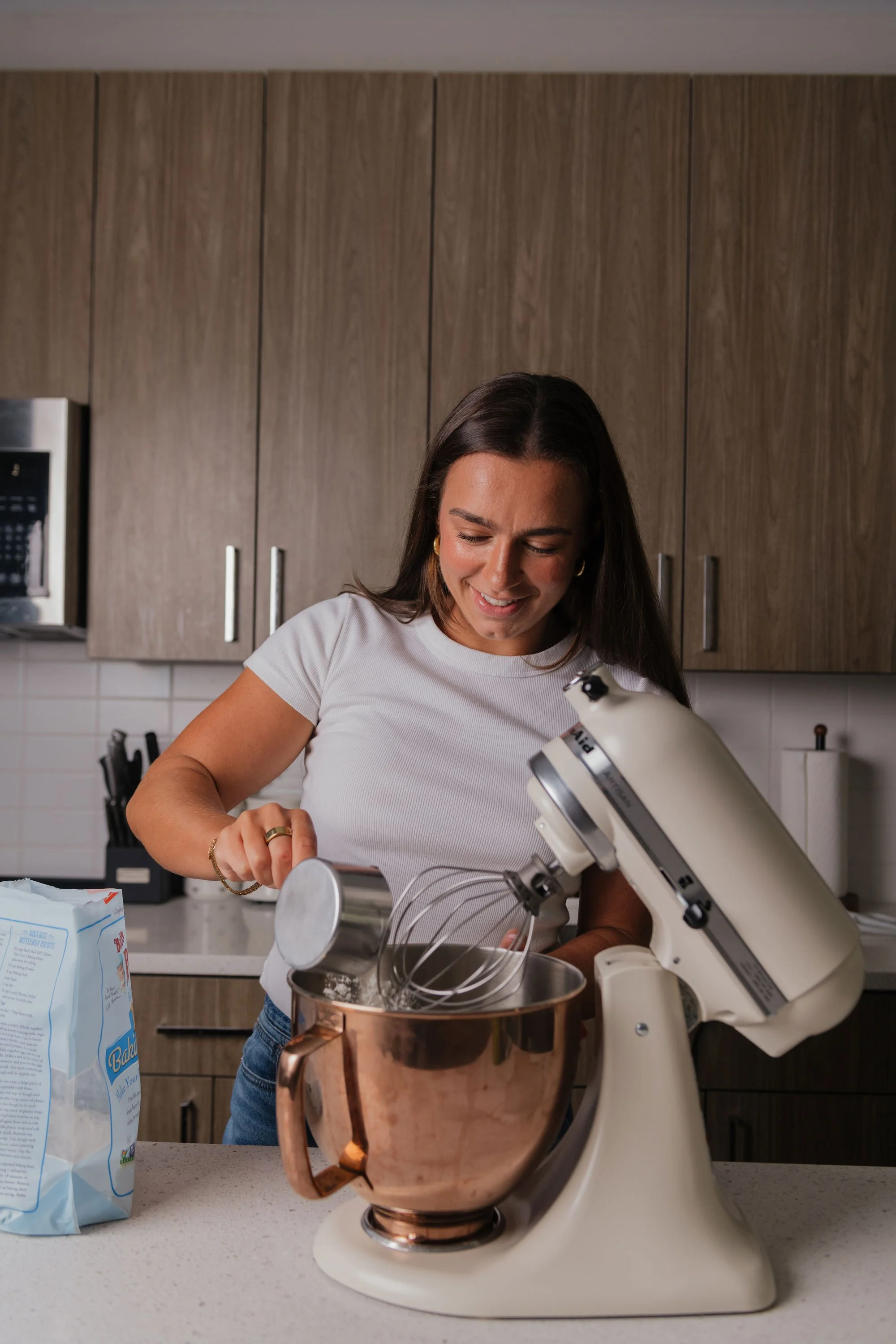 A woman smiling and baking with a stand mixer in a modern kitchen.