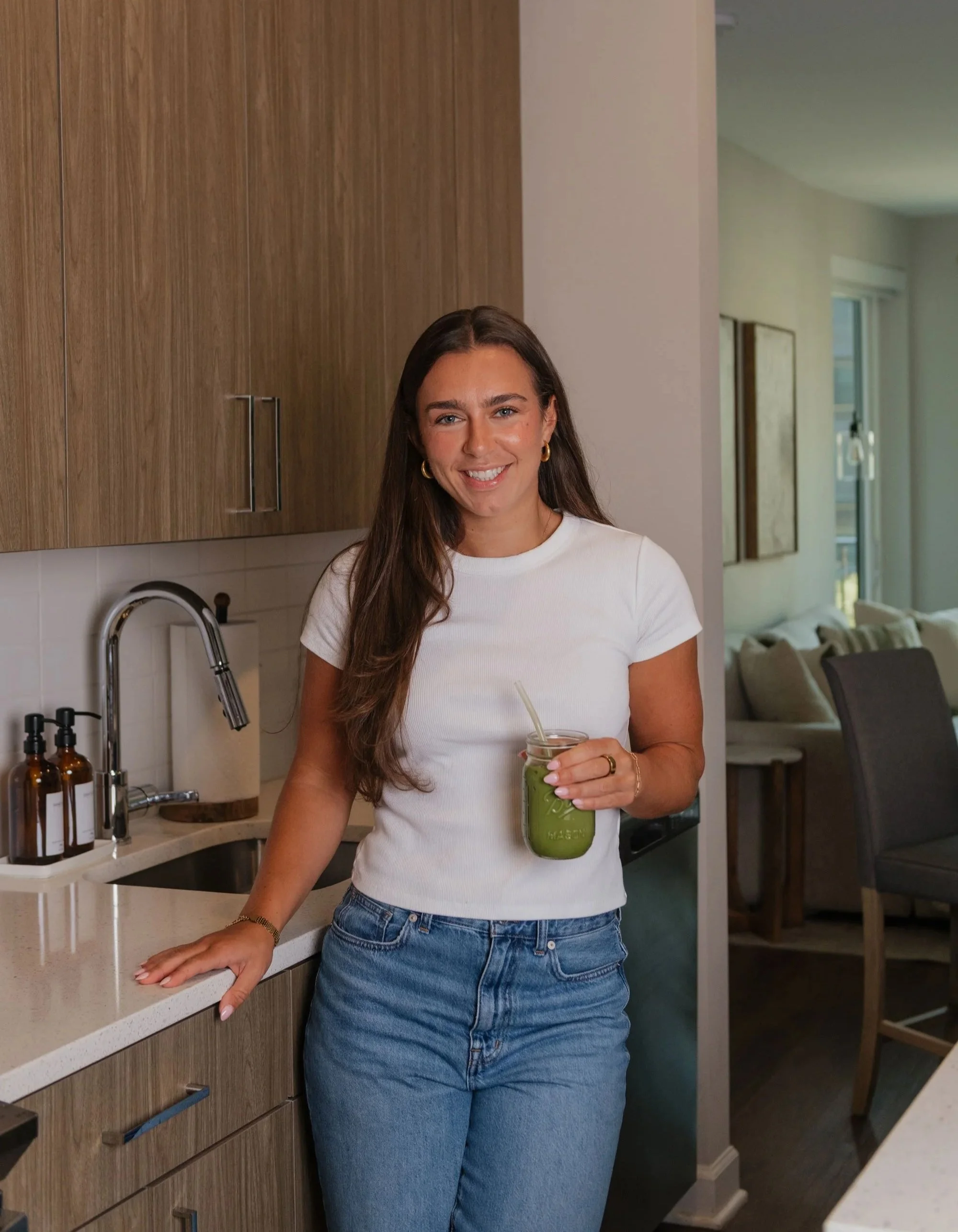 A woman with long brown hair, wearing a white t-shirt and blue jeans, standing in a modern kitchen holding a glass jar of green smoothie with a straw, smiling at the camera.