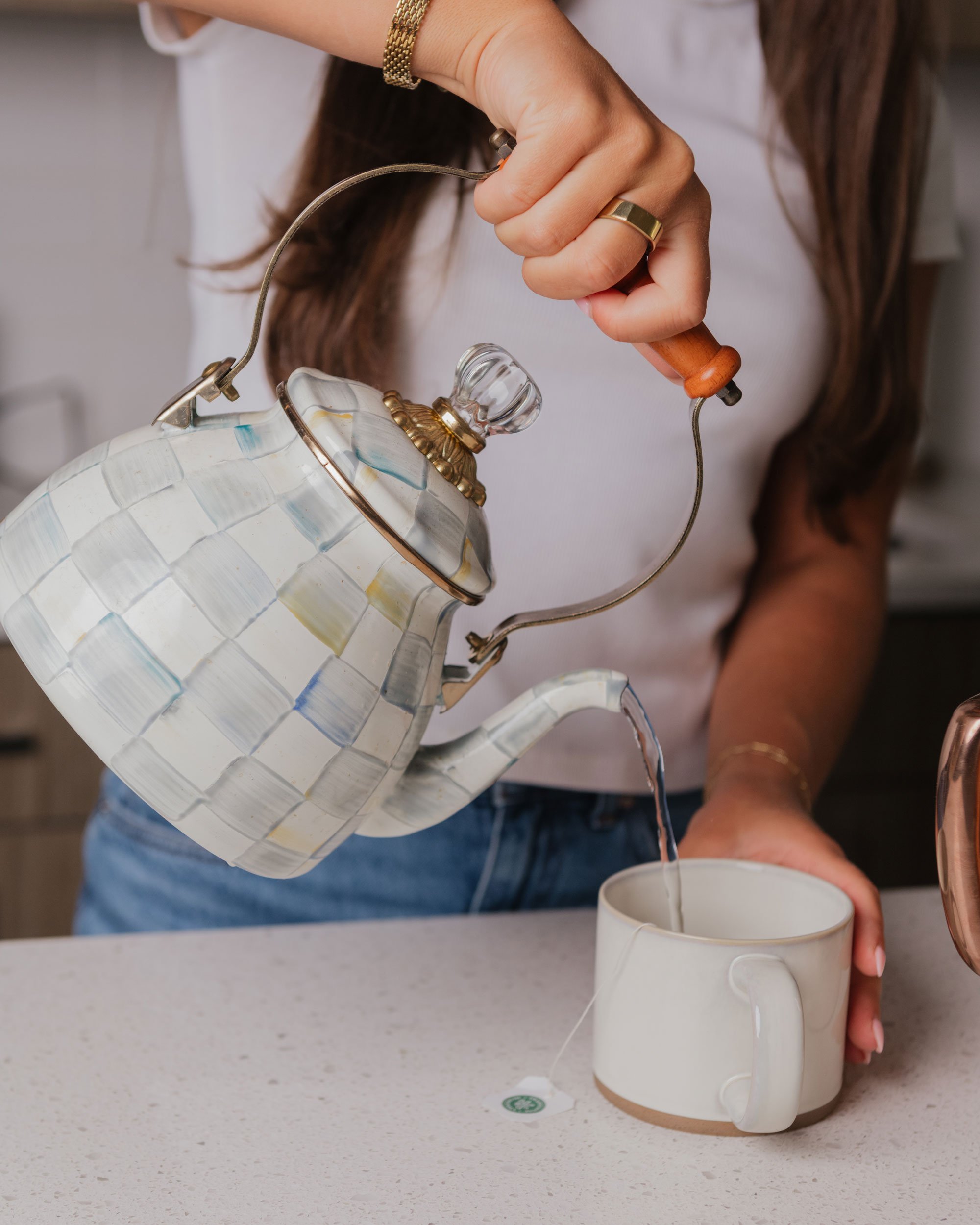 Person pouring hot water from a decorative ceramic teapot into a ceramic mug on a kitchen countertop.