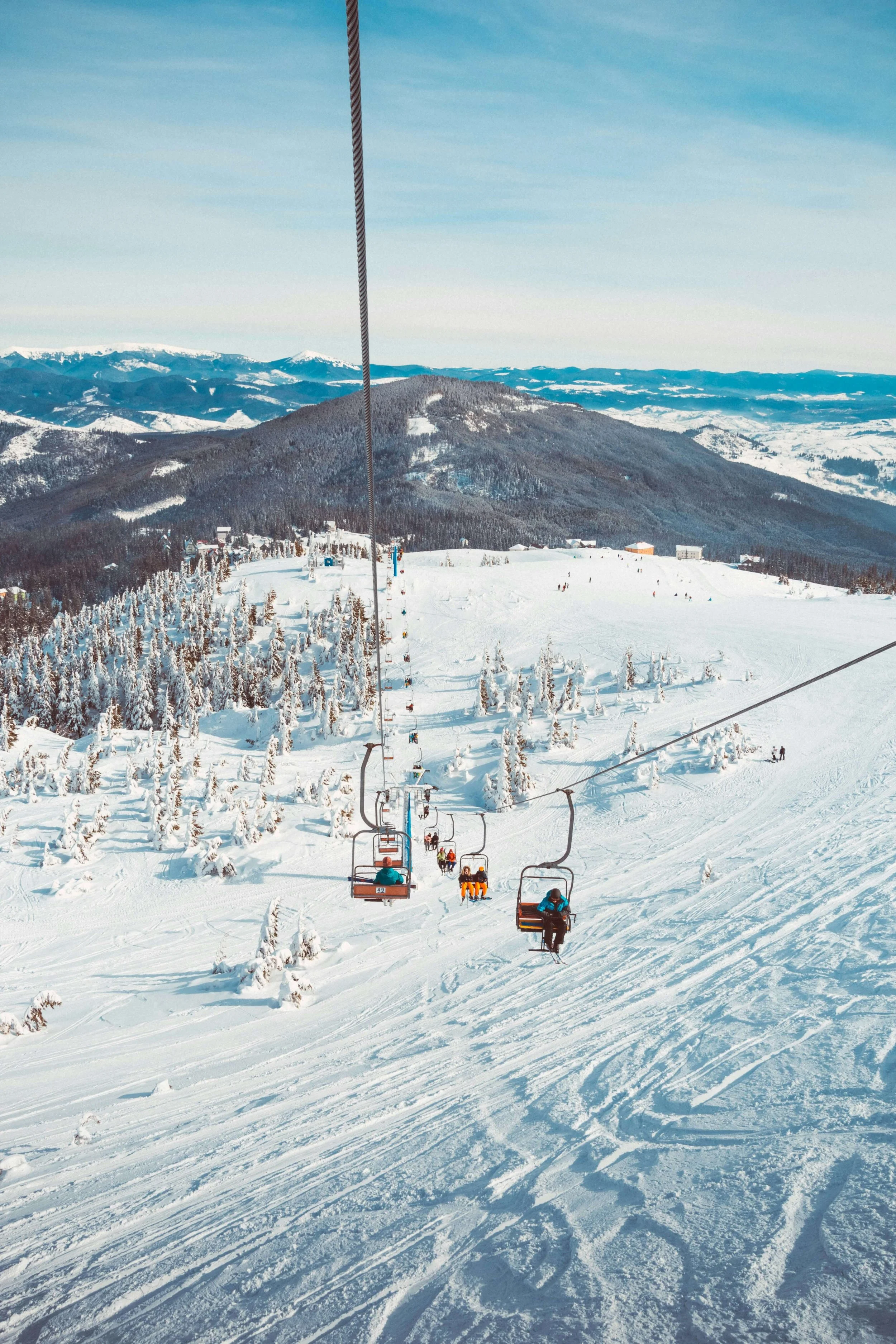 Snow-covered ski slopes with chairlifts and skiers, surrounded by snow-laden trees and mountain ranges in the distance.
