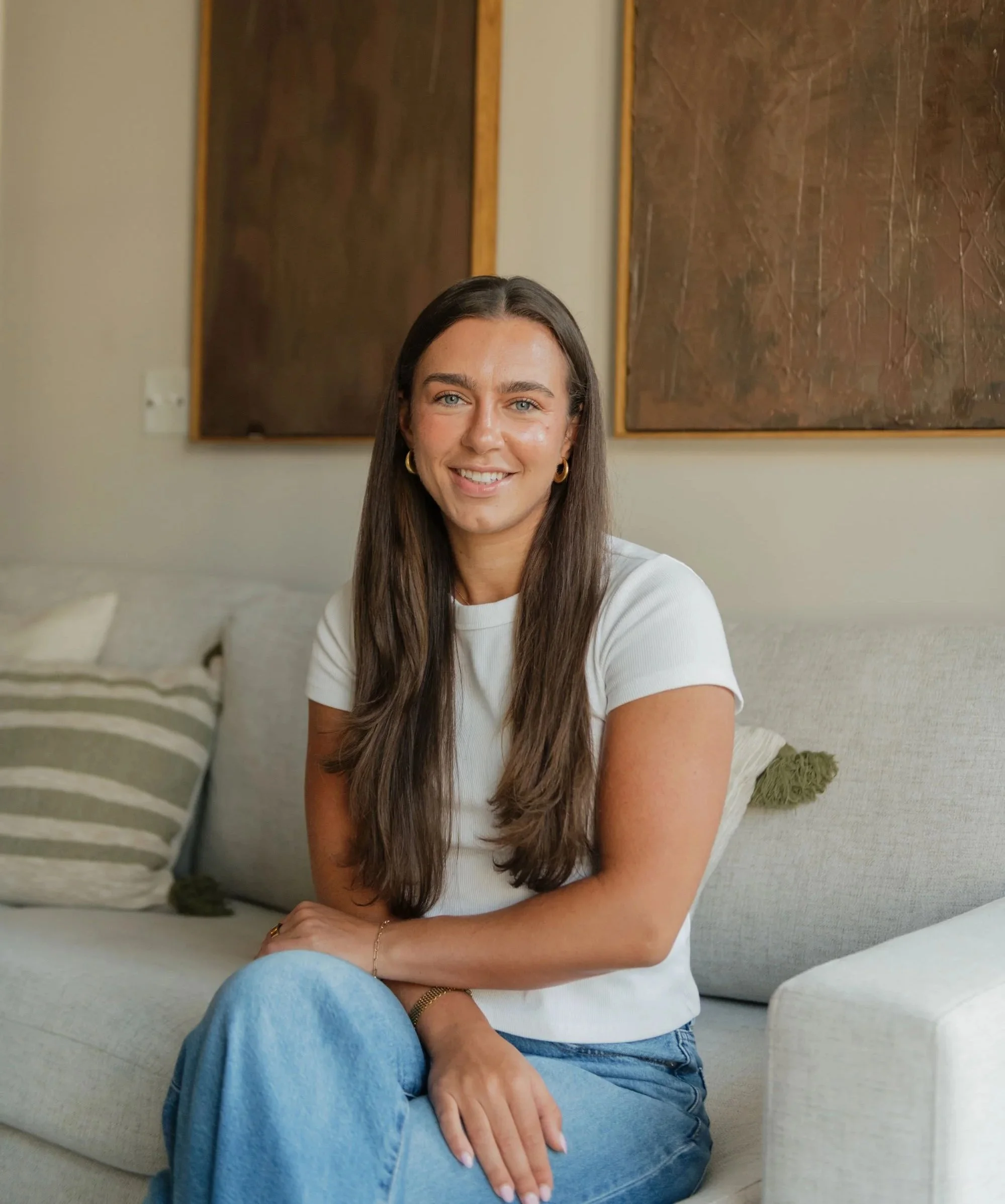 A woman with long brown hair, wearing a white t-shirt and blue jeans, sitting on a beige couch in a room with abstract paintings on the wall behind her, smiling at the camera.