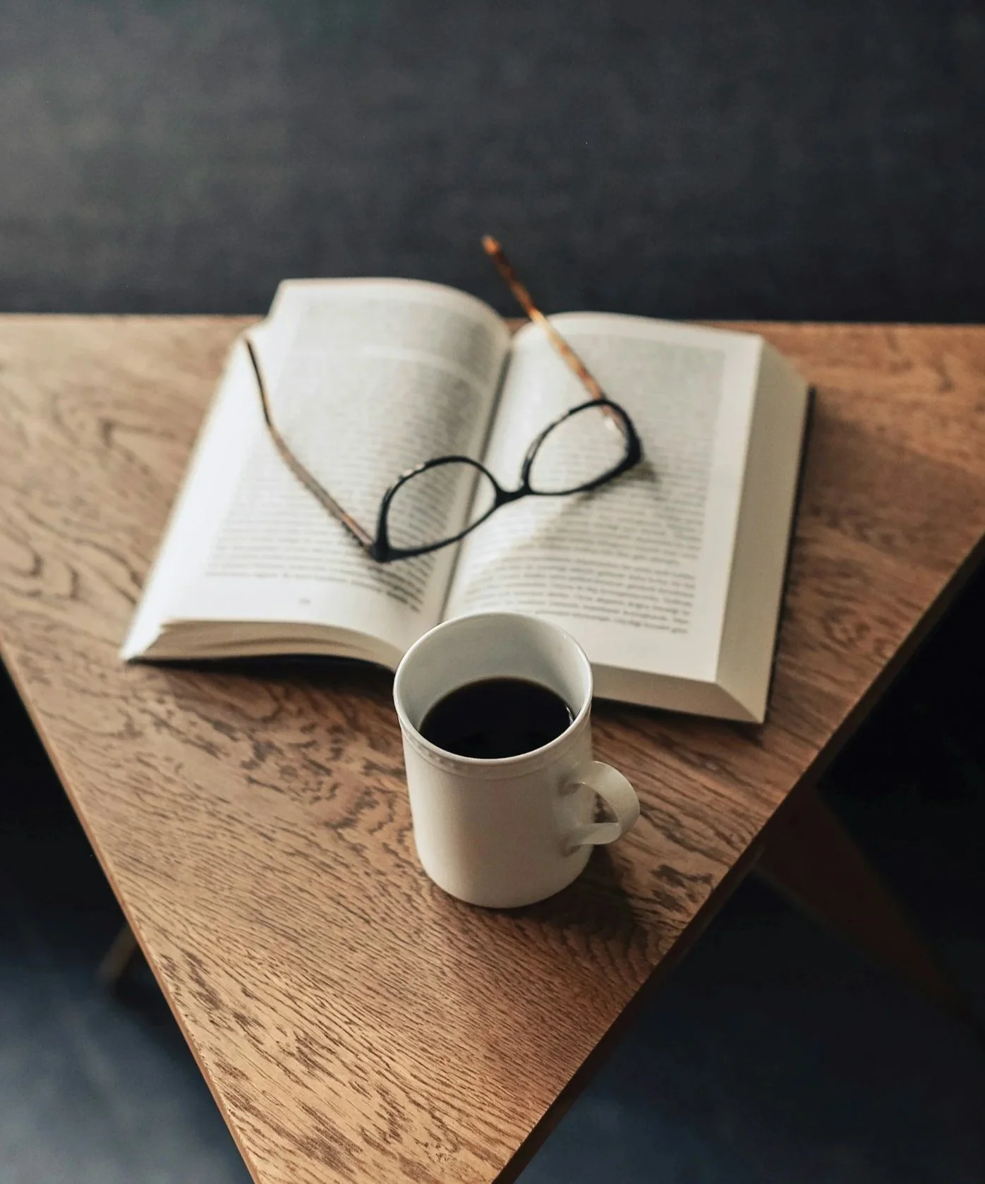 An open book with a pair of glasses on top, a cup of black coffee in front, and a wooden table with a dark background.