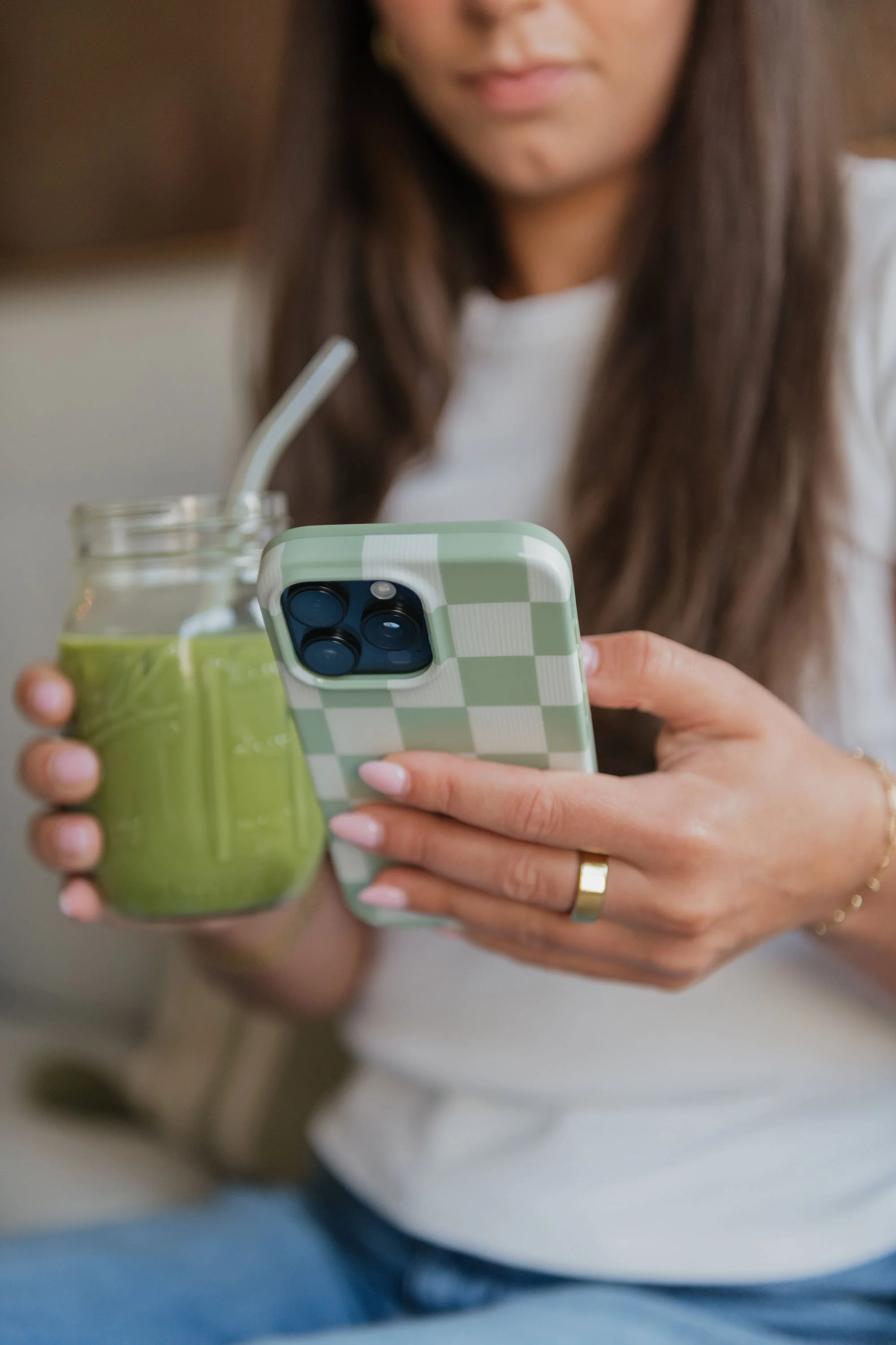 A woman holding a mason jar with a green smoothie and a straw, looking at her smartphone with a green and white checkered case.