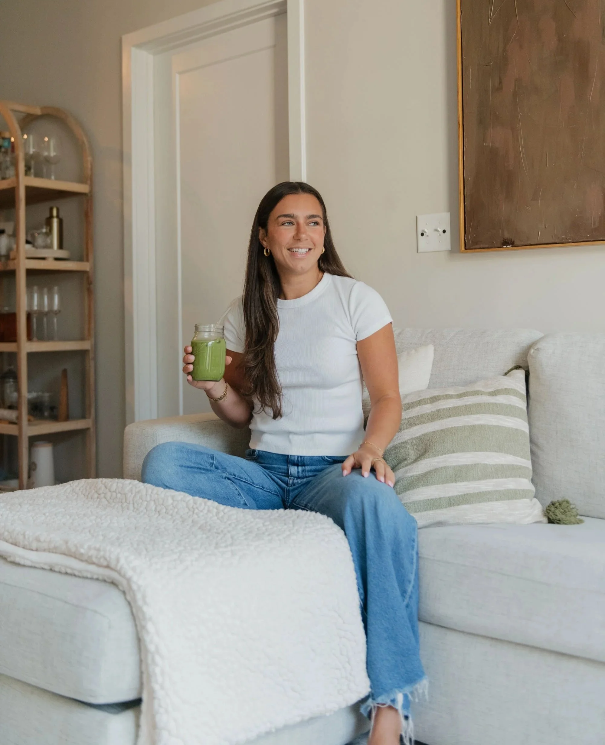 A young woman with long dark hair, wearing a white t-shirt and blue jeans, sitting on a light-colored sofa with striped pillows, holding a green smoothie jar, smiling indoors.