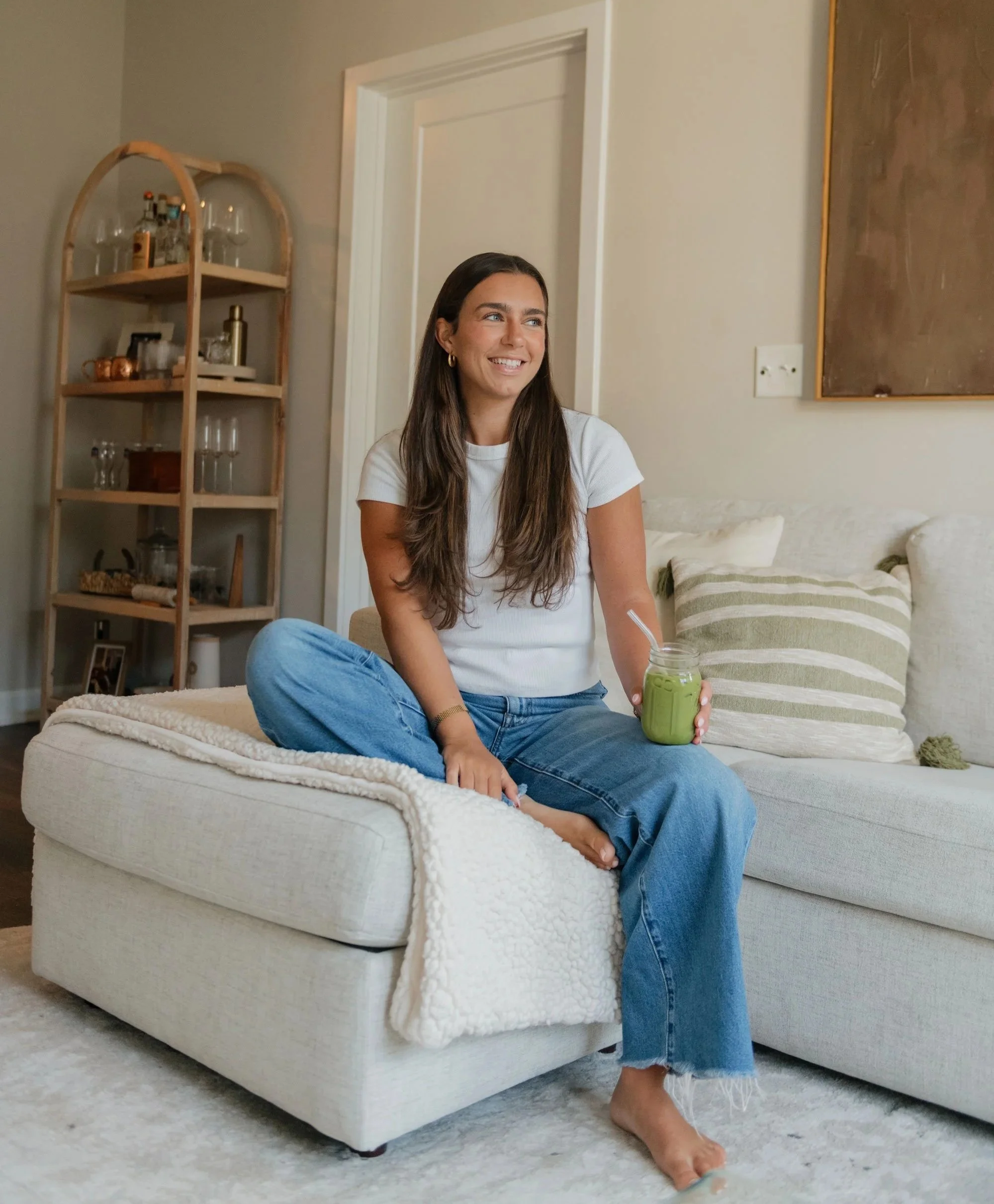 A woman sitting cross-legged on a white couch in a living room, holding a green smoothie with a straw, smiling. There is a beige and brown striped pillow on the couch and a beige blanket. In the background, a wooden shelf with glassware and decorative items, and a door.