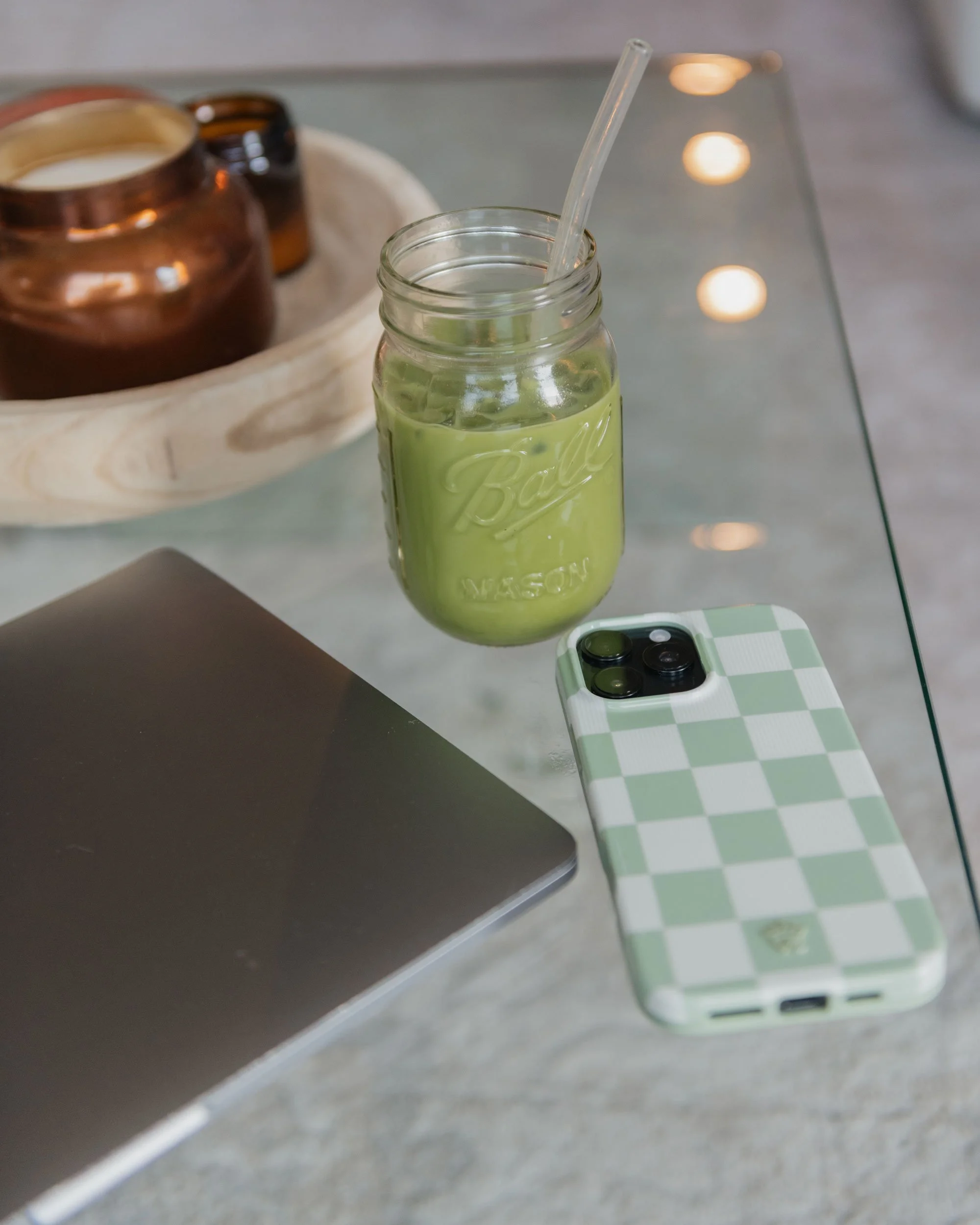 A glass jar with a green drink and a straw, a smartphone with a green and white checkered case, a closed laptop, and a wooden tray with brown jars on it, on a glass table.