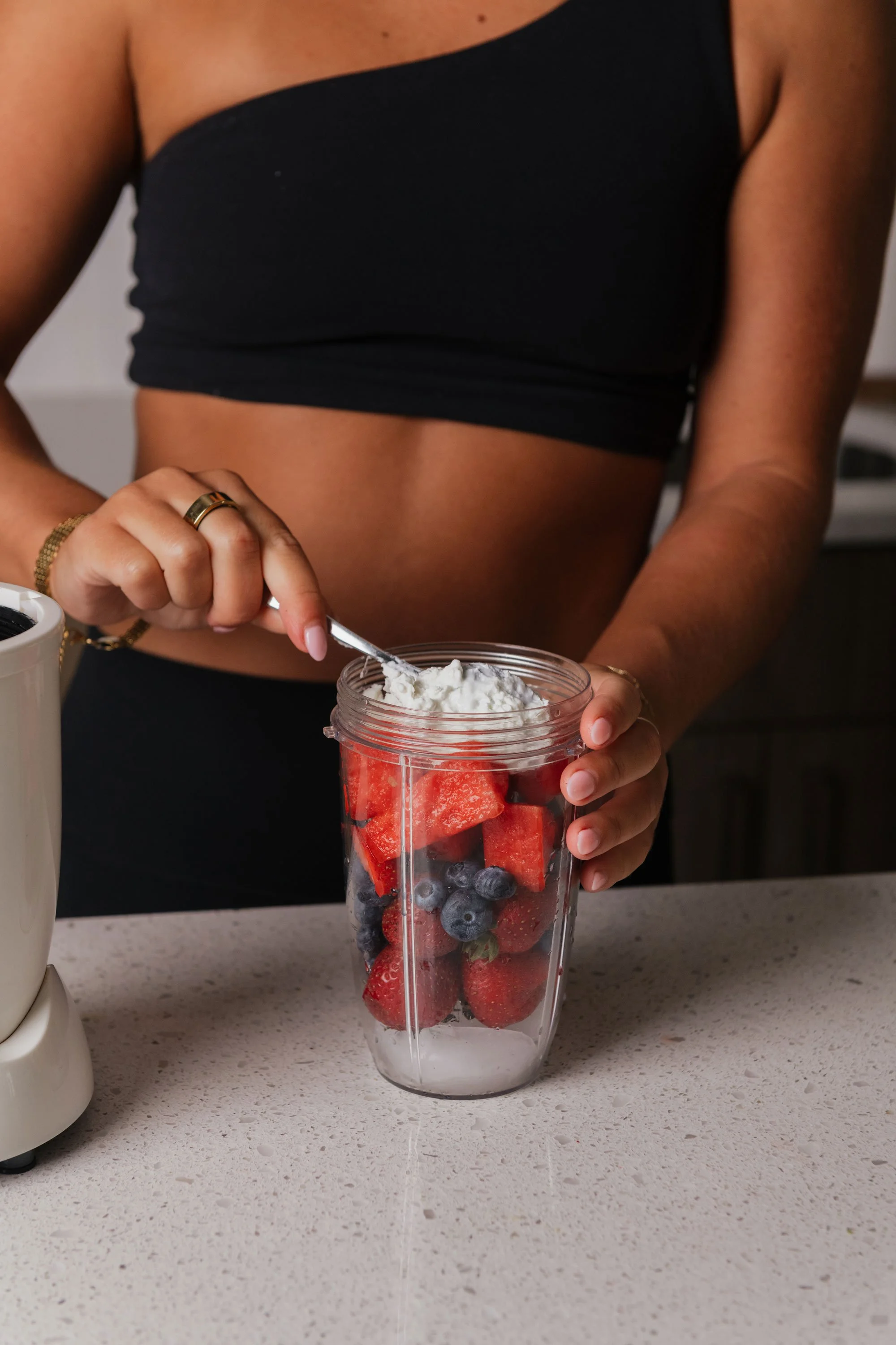 A woman in a black tube top preparing a smoothie with strawberries, blueberries, and watermelon in a blender, adding whipped cream on top.