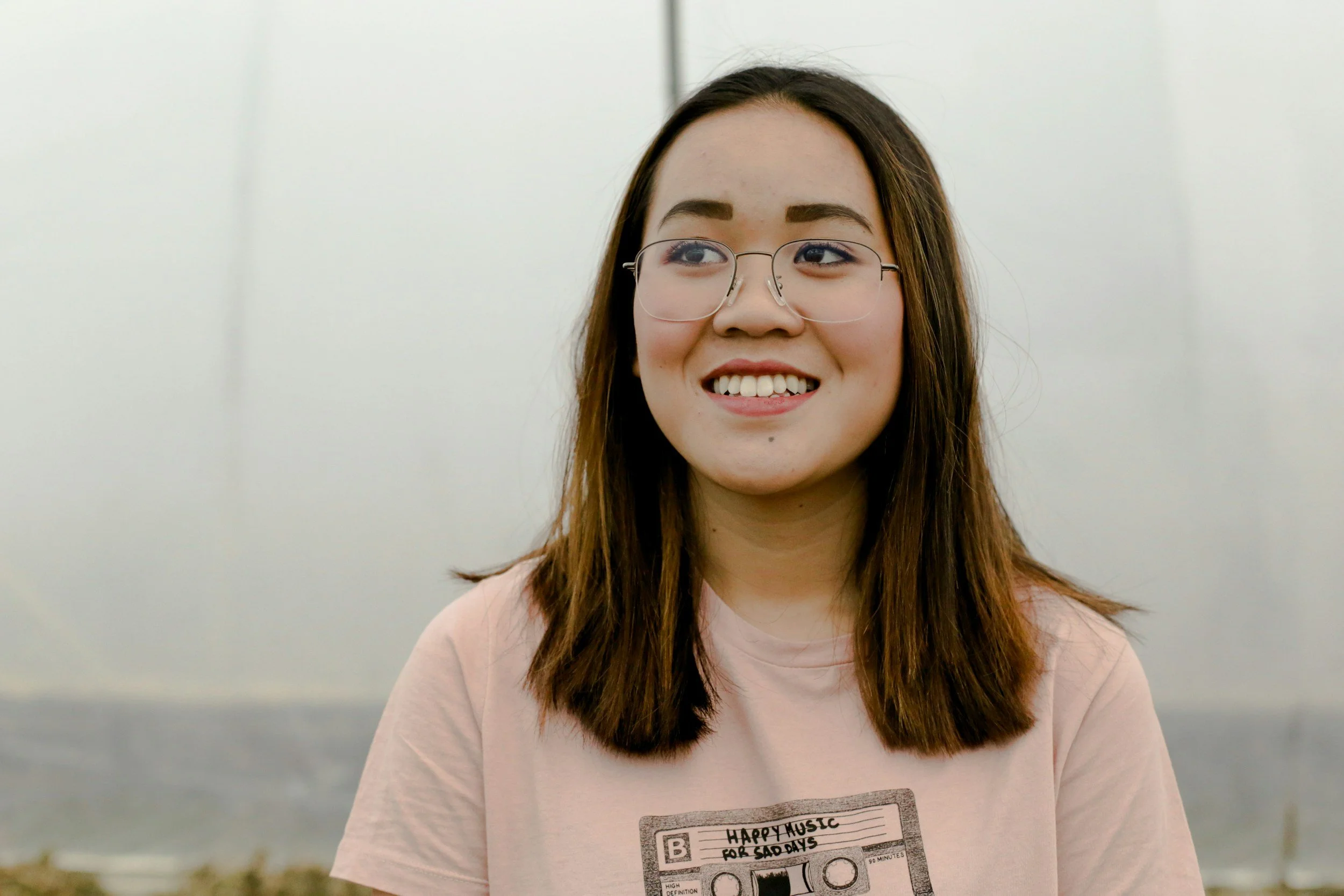 A young woman with long brown hair, glasses, and a light pink T-shirt smiling outdoors.