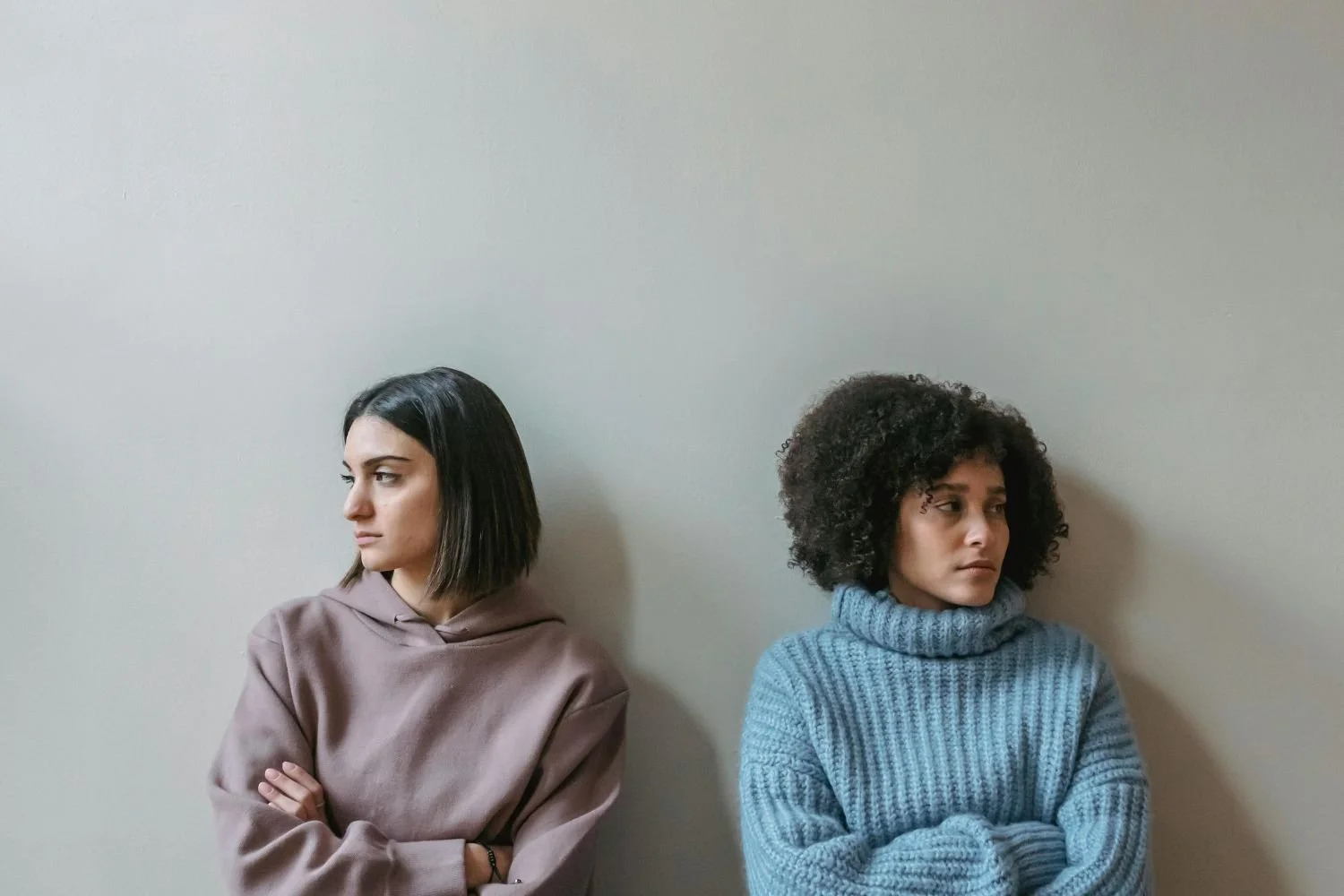Two women standing against a plain wall, arms crossed, looking away from each other with serious expressions.