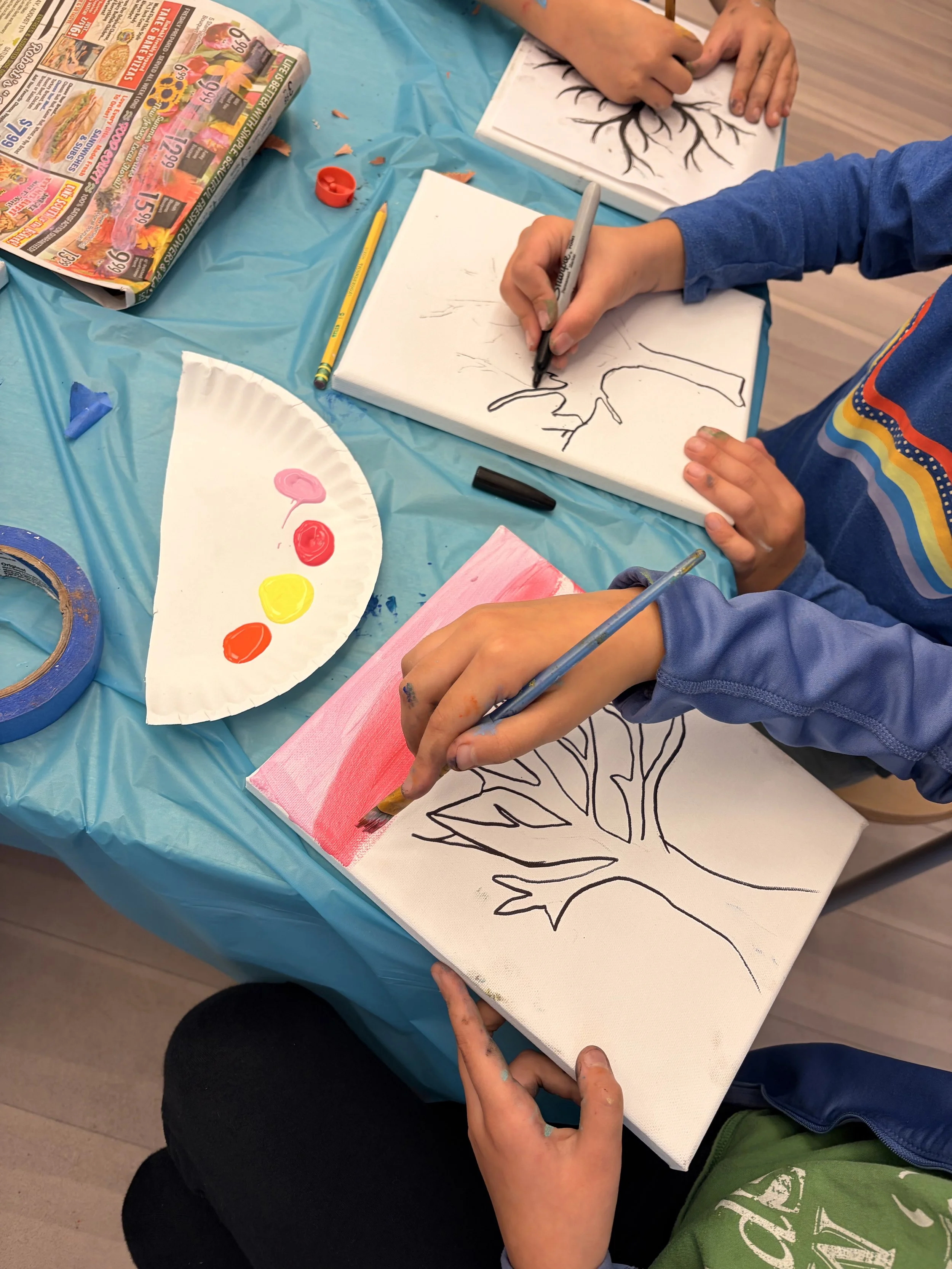 Children painting and drawing pictures of trees on canvases during an art activity. One canvas shows an outline of a tree, and a child is adding colors to a section using a paintbrush. Various art supplies are on the table, including a paint palette, pencils, and tape.