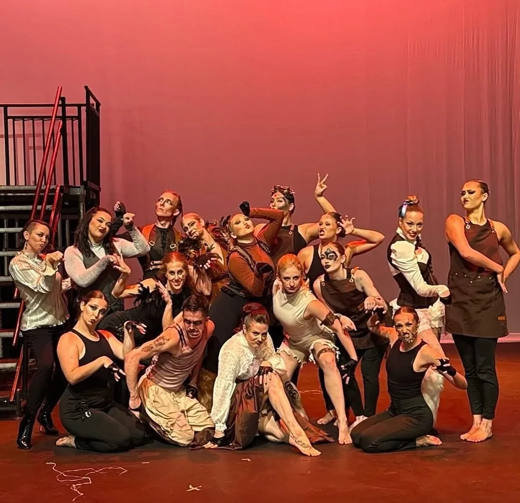 Group of performers in theatrical costumes on stage posing for a photo with expressive gestures and facial expressions.