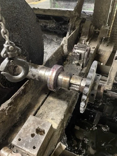 Close-up of industrial machinery, including a metal shaft, gears, and a chain attached to a large metal component used in manufacturing or machining.
