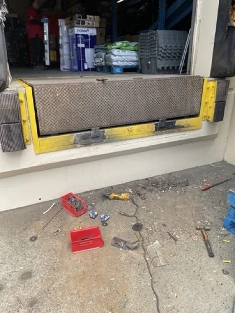 A loading dock with a yellow bumper guard, open for shipping or receiving. Tools and small items scattered on the concrete ground include a tape measure, a drill, and a red toolbox. In the background, shelves with various supplies and containers are