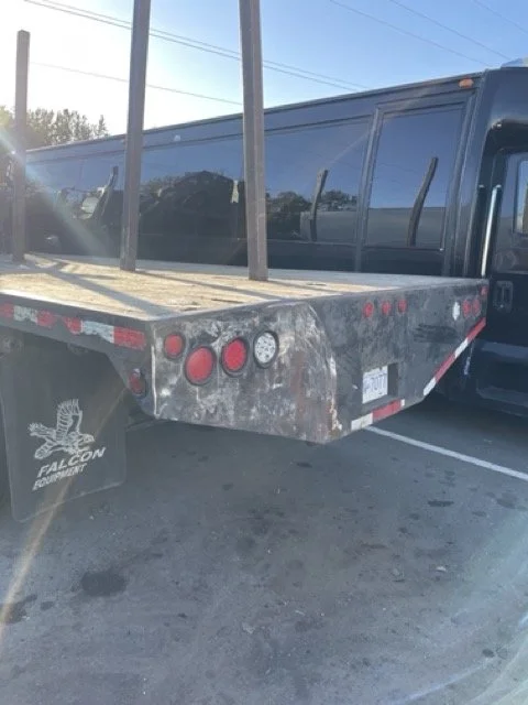 Flatbed truck with a dark-colored cab, parked in a lot with power lines above, and some trees in the background.