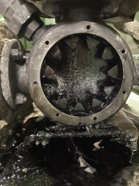 Close-up of a mechanical component, possibly part of a machine or engine, with dark oil or grease inside, surrounded by a metal frame, in a workshop or industrial setting.