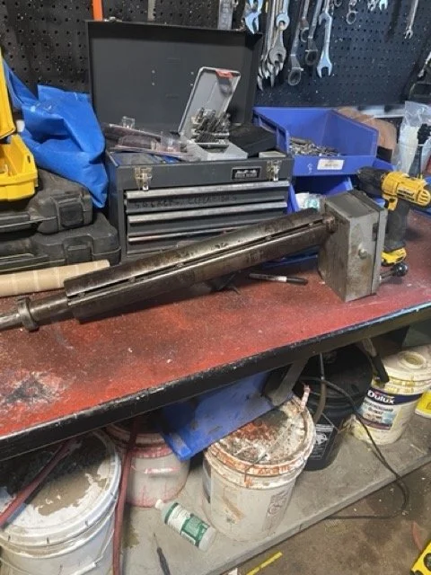 Workbench with a large metal shaft, a toolbox with various tools inside, and a yellow cordless drill, with buckets and miscellaneous items underneath.