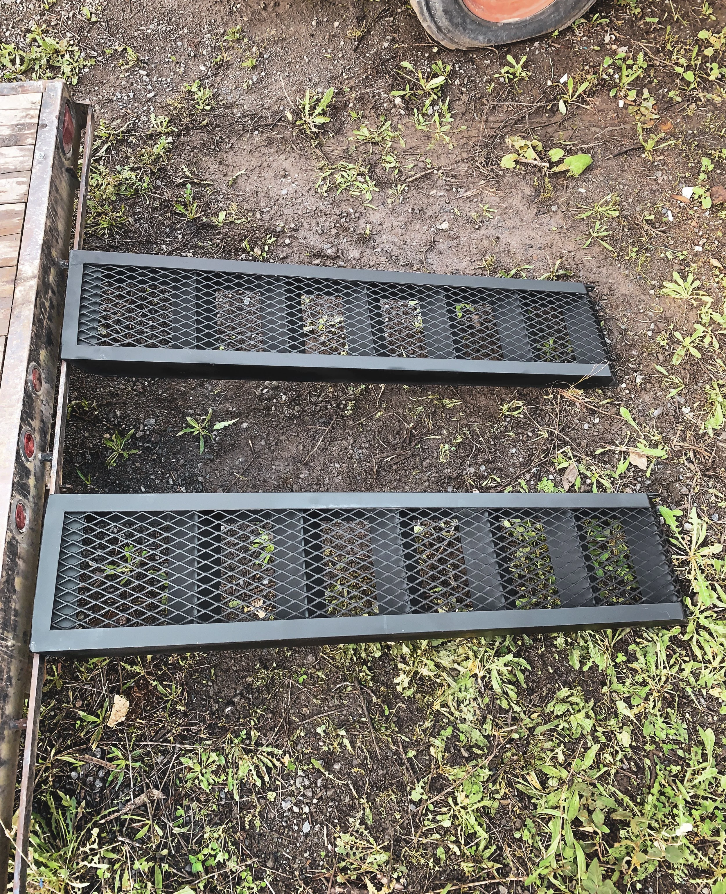 Two black metal grates with diamond pattern on a patch of dirt and grass, with a trailer on the left and a wheel of a tire in the upper right corner.