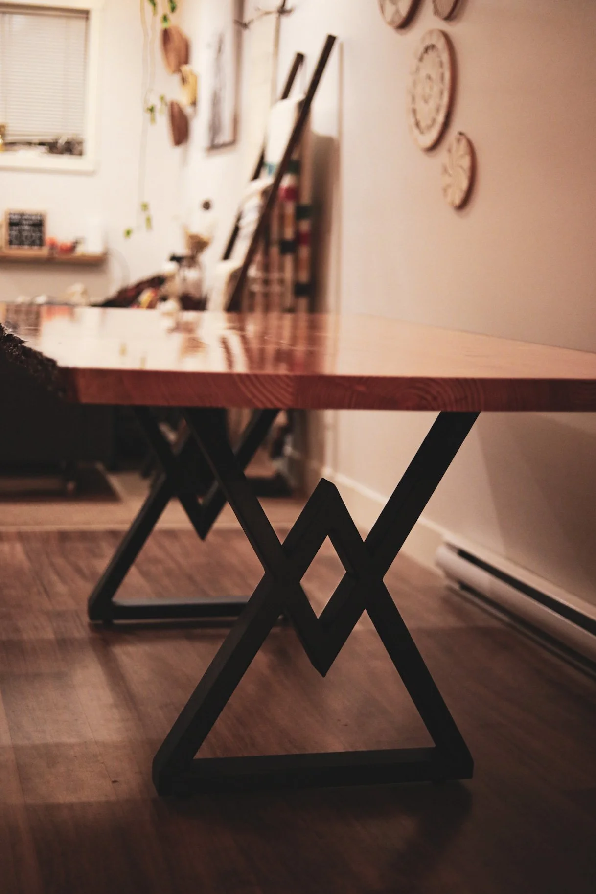 Close-up view of a wooden table with a black metal geometric base in a room with wall decor and a window in the background.