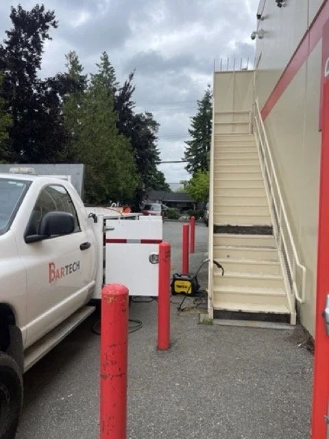 A white pickup truck parked next to a set of outdoor stairs leading to a building entrance, with red bollards in the parking lot and trees in the background.