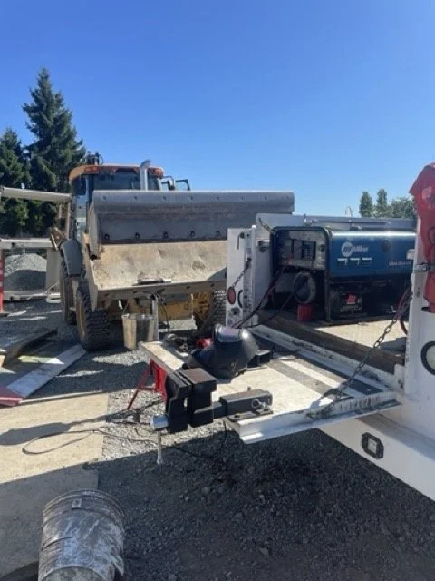 Construction site with a large snow plow attached to a pickup truck, a generator, and various tools on a trailer, under a clear blue sky.