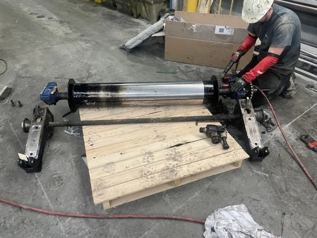 A worker using a power tool on a large hydraulic piston mounted on a wooden pallet in a workshop.