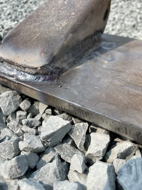 Close-up of a welded metal joint on a steel plate resting on gravel.