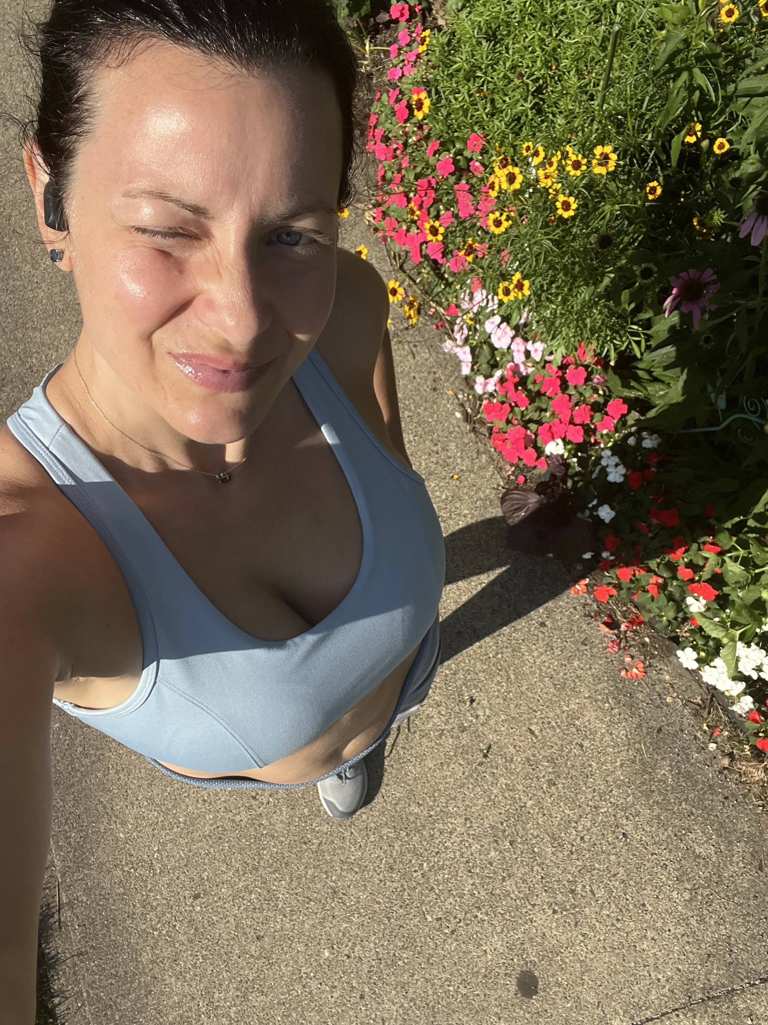Denise Maloney taking a selfie on a sidewalk next to a colorful flower bed of pink, yellow, red, purple, and white flowers in sunlight.