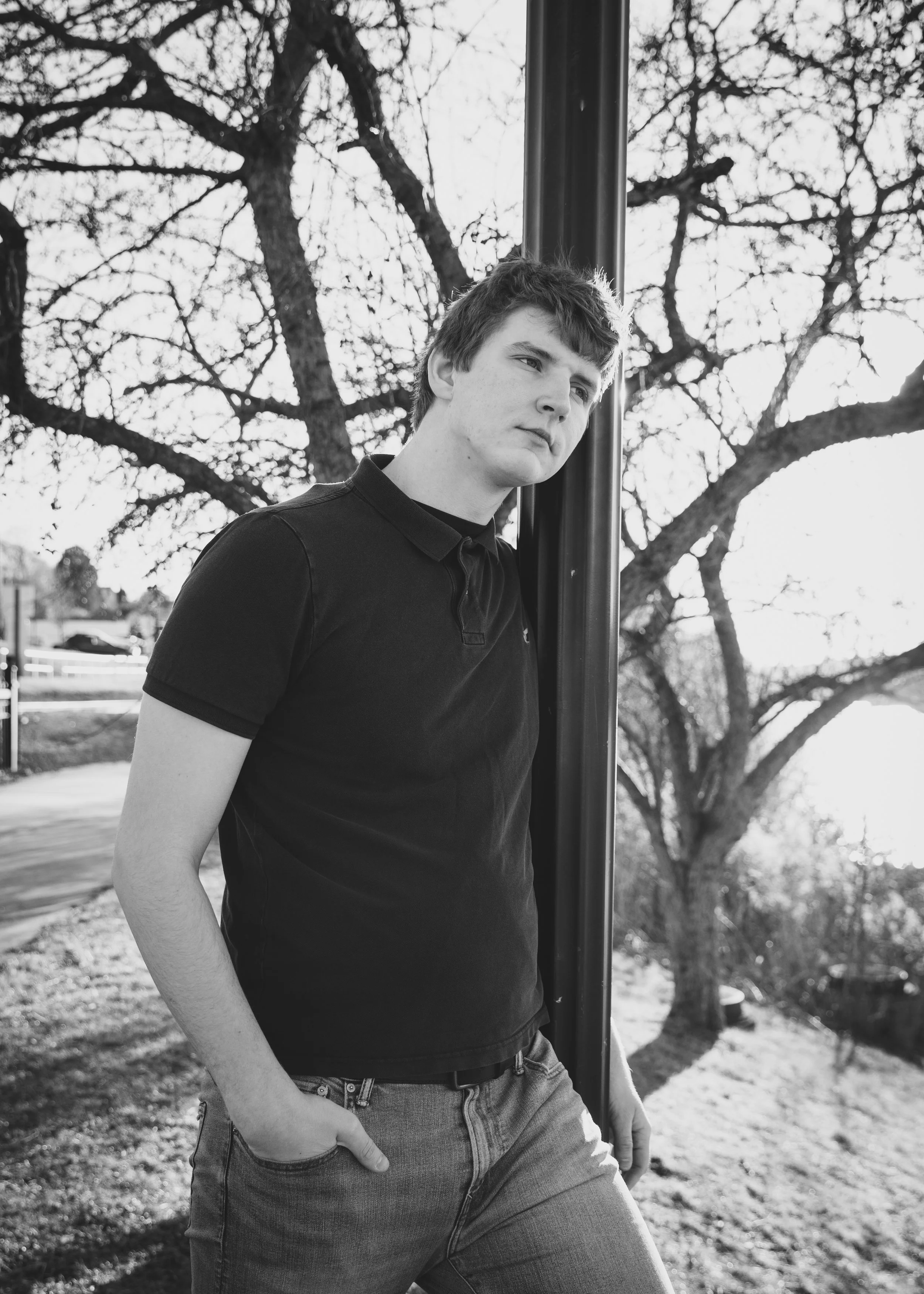 A young man leaning against a pole outdoors in a park with trees in the background, wearing a black polo shirt and jeans, looking to the side.