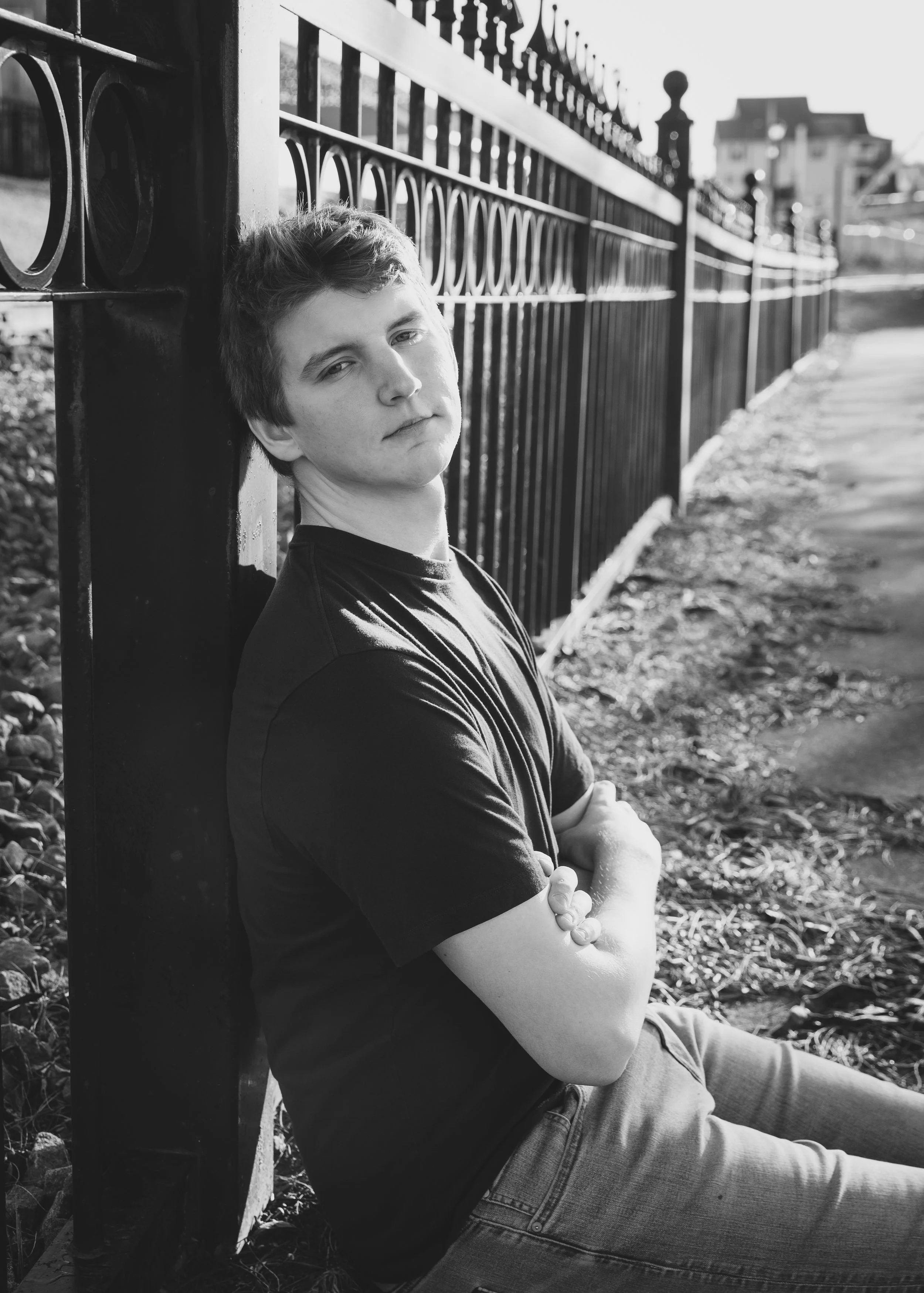 A young man with short hair and a serious expression sitting against a black metal fence, arms crossed, outdoor setting with grass and a sidewalk.