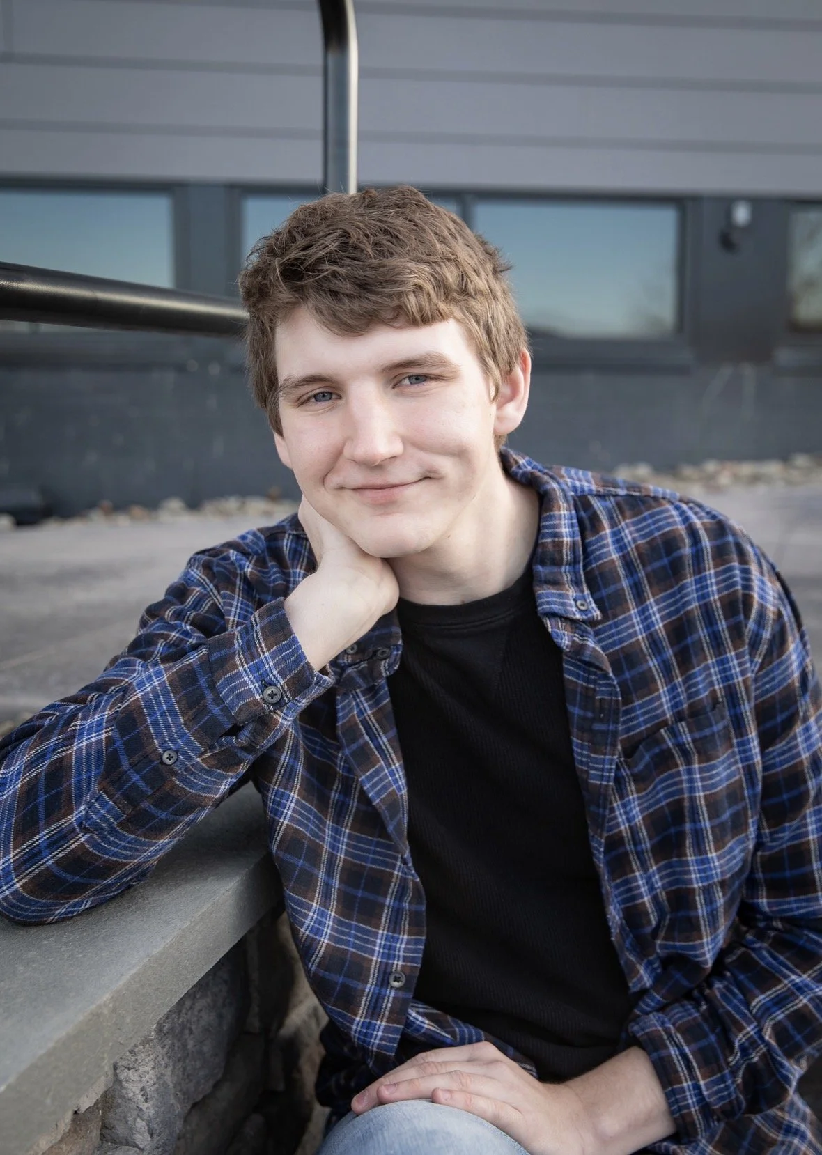 Young man in plaid shirt sitting outdoors, resting his chin on his hand, smiling softly, with modern building in the background.
